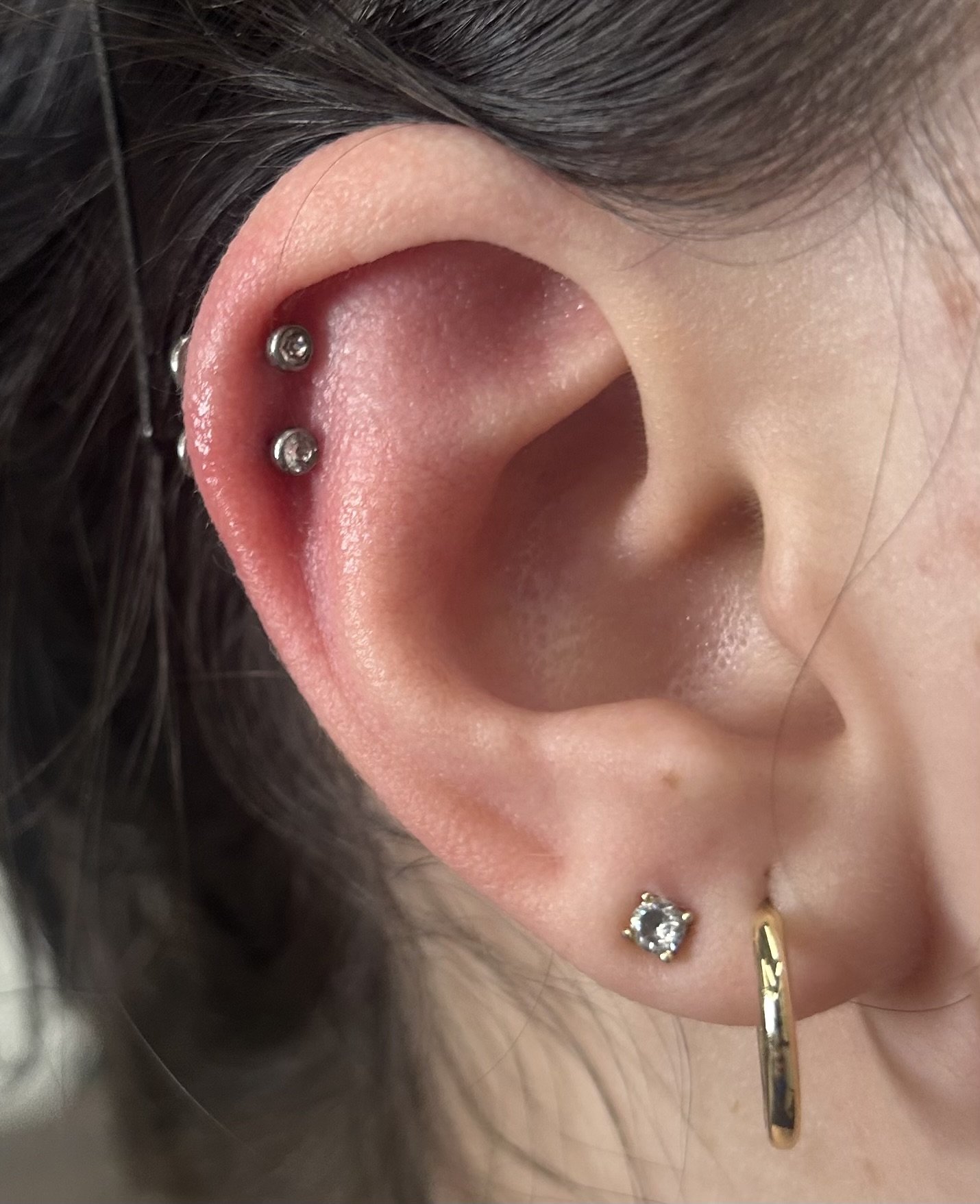 Close-up of a human ear with multiple piercings, including two silver balls in the upper cartilage, a stud with a clear gemstone in the earlobe, and a gold hoop earring.