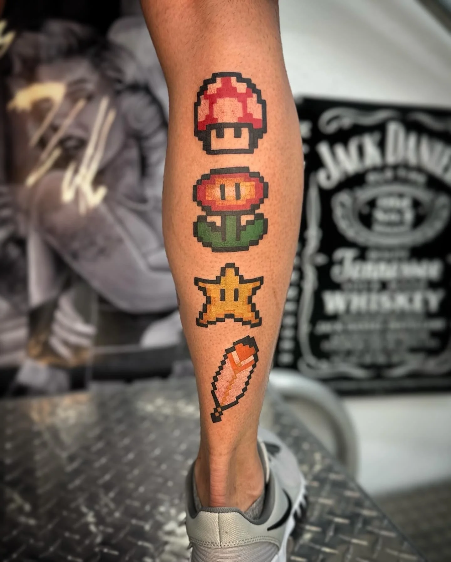 Colorful pixel art tattoo on lower leg featuring a Super Mushroom, a Fire Flower, a Super Star, and a Fire Stick from Mario video game.