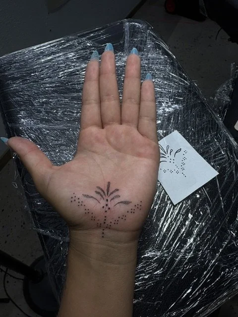 A person's left hand with a temporary tattoo of a firework design on the palm, resting on a clear plastic-covered surface, with a small decorated card nearby.