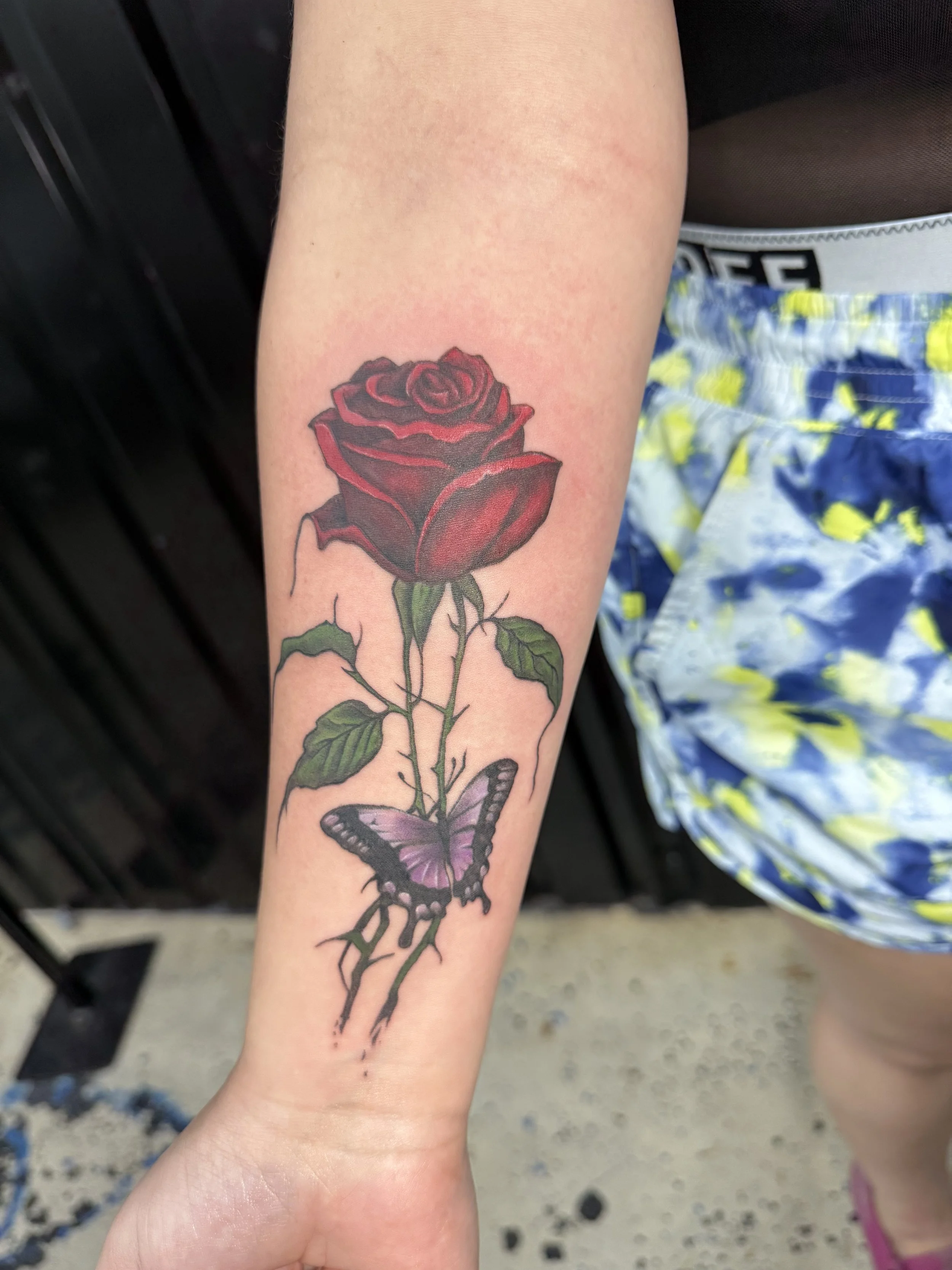 Tattoo of a red rose with green leaves and a purple butterfly on the person's inner forearm.