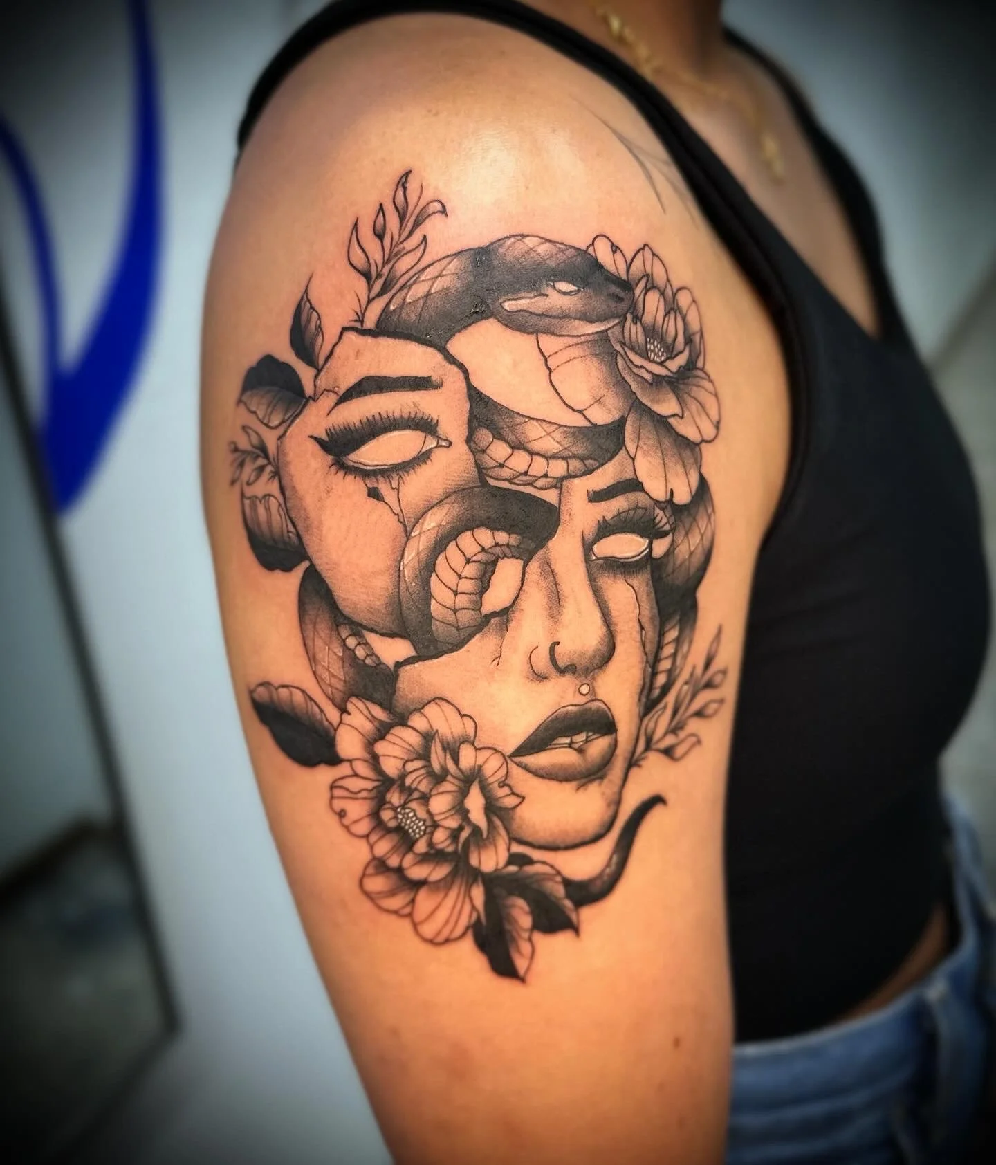 Black and white tattoo of a woman's face with closed eyes, tears, a snake, and flowers on upper arm.