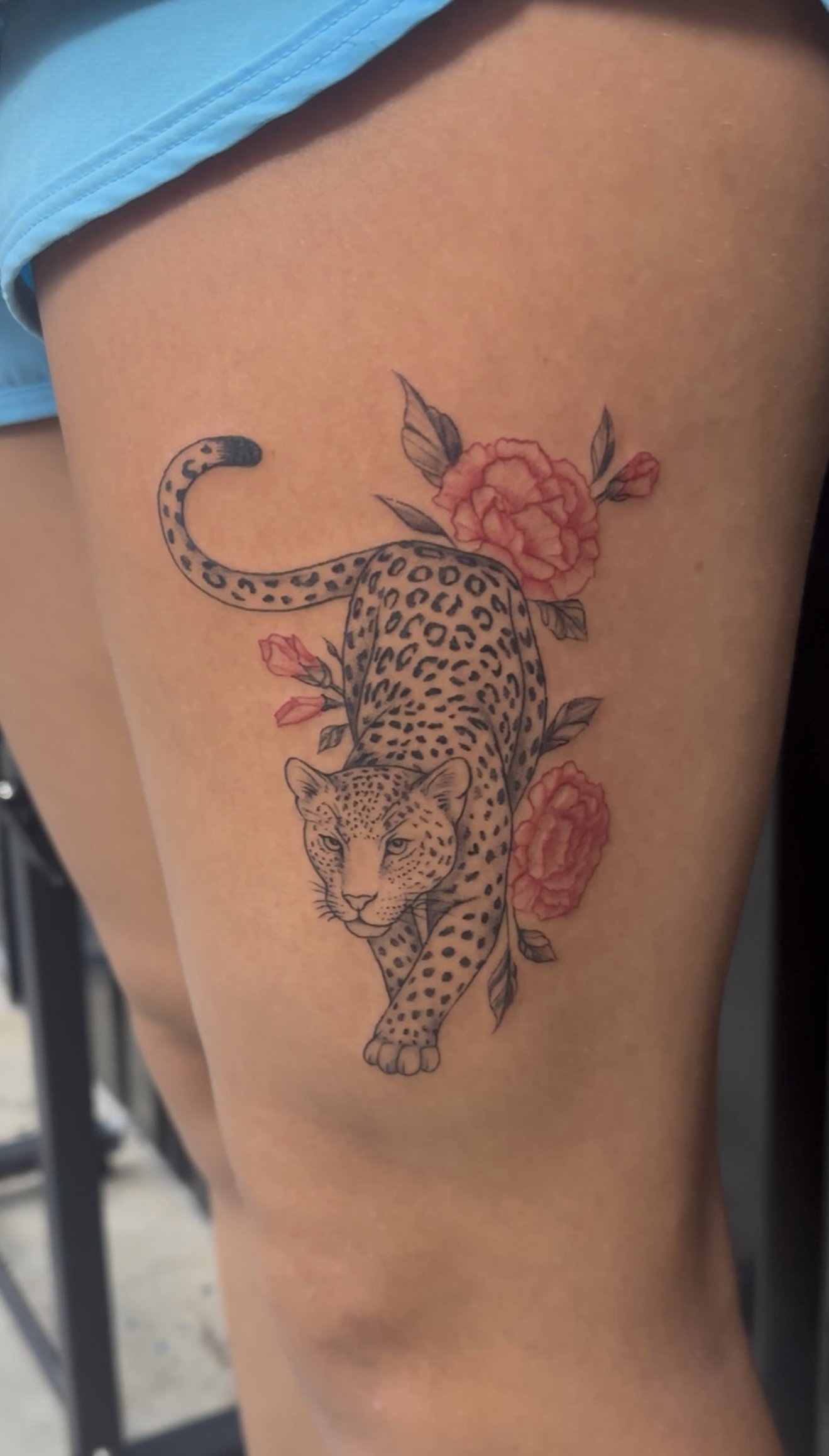 Tattoo of a leopard walking among pink flowers and green leaves on a person's thigh.
