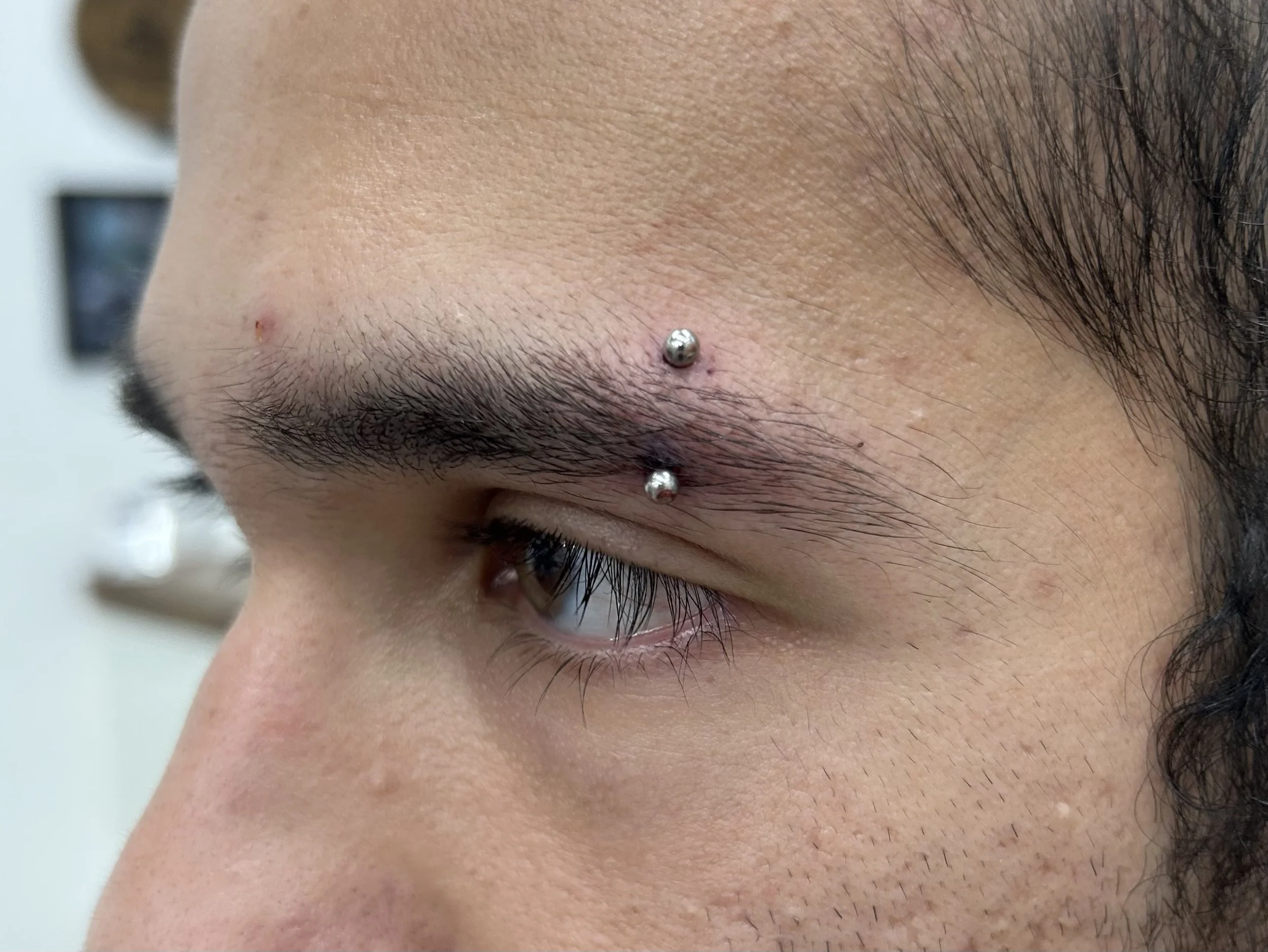 Close-up of a person's forehead showing a nose piercing with a shiny silver ball and a small red blemish or pimple nearby.