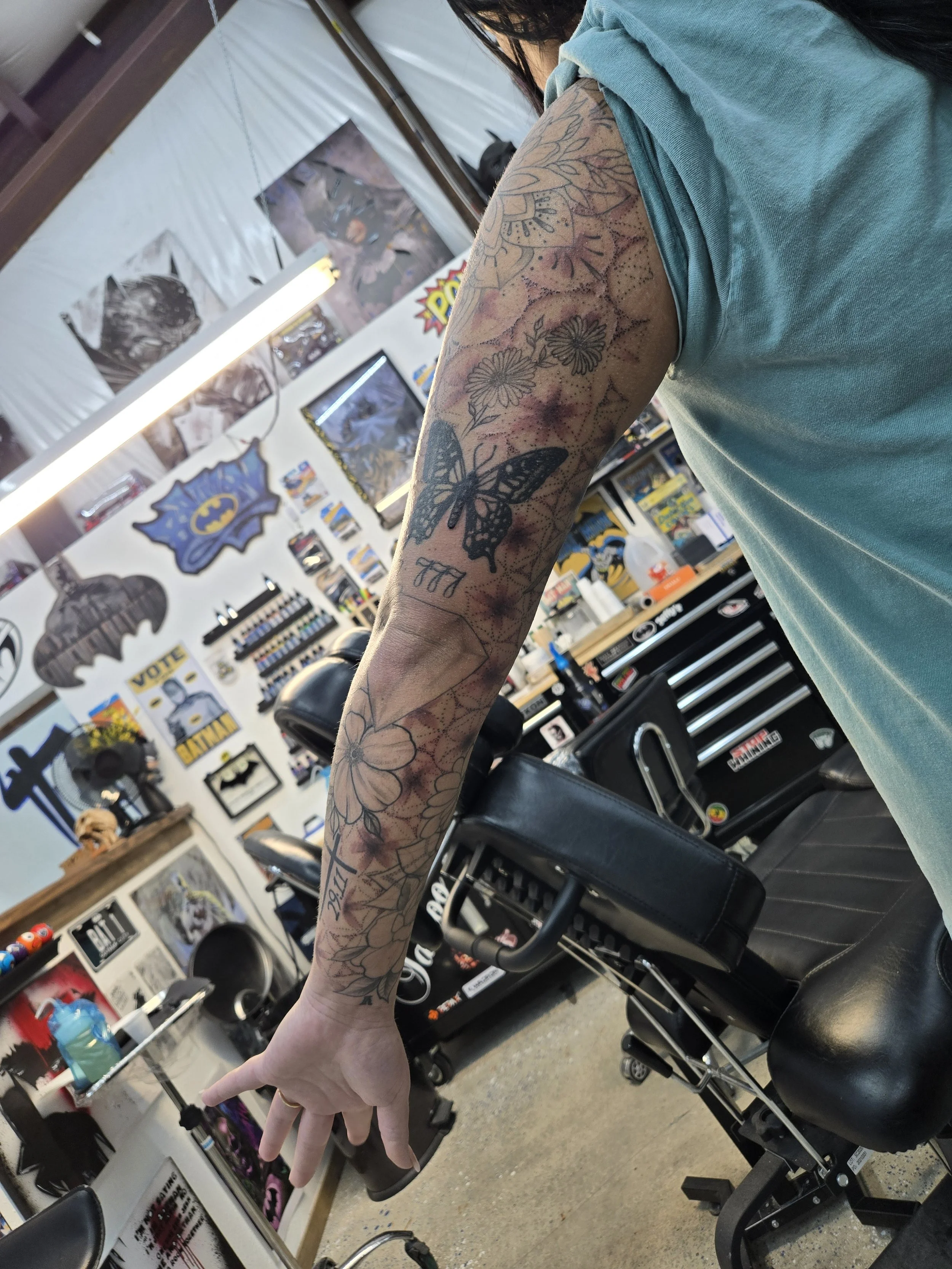 Tattooed arm of a person in a tattoo shop. Visible tattoos include a butterfly, flowers, and geometric patterns.