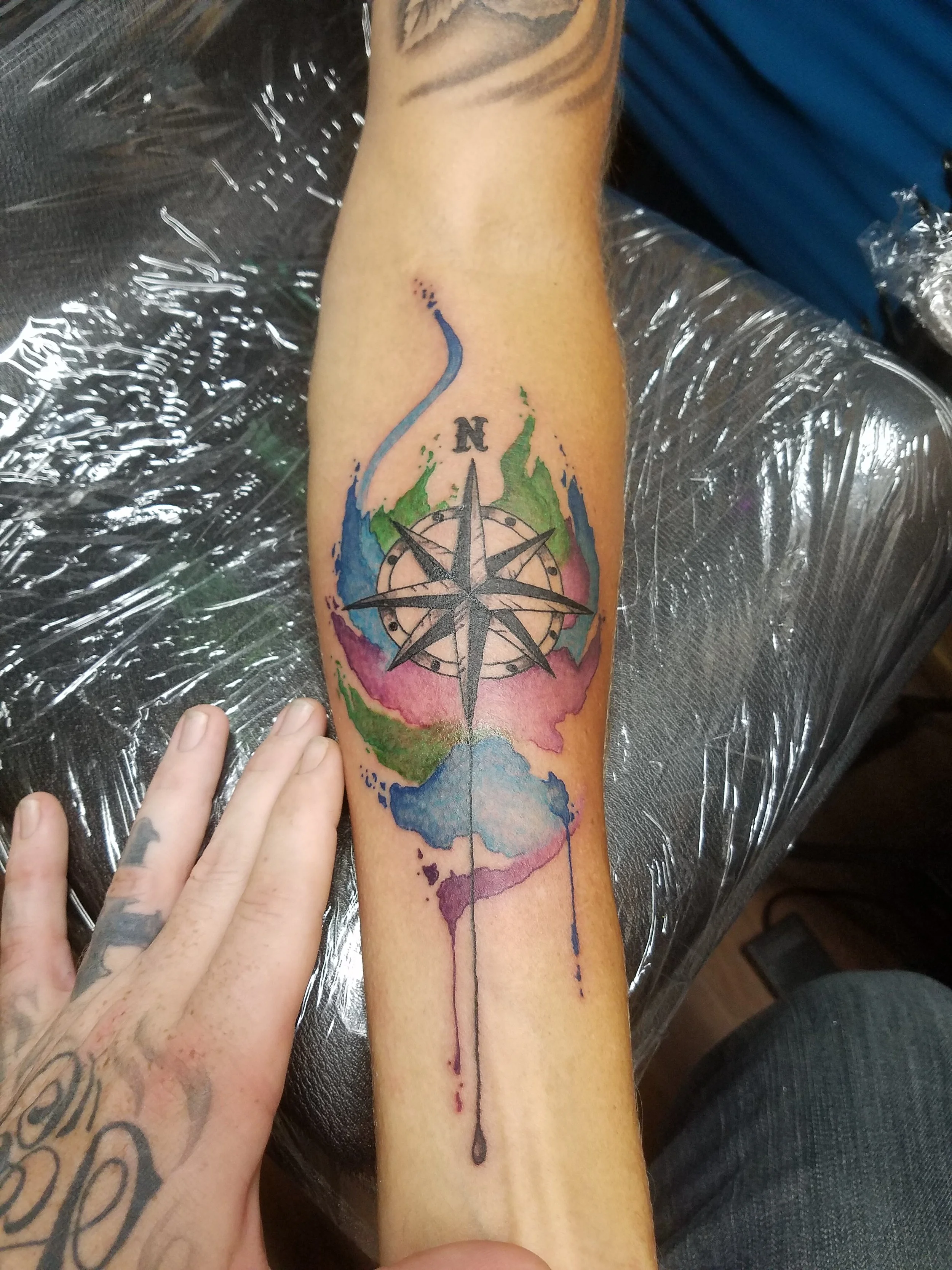 Colorful watercolour tattoo of a compass with a north needle, surrounded by abstract paint splashes in green, pink, blue, and purple.