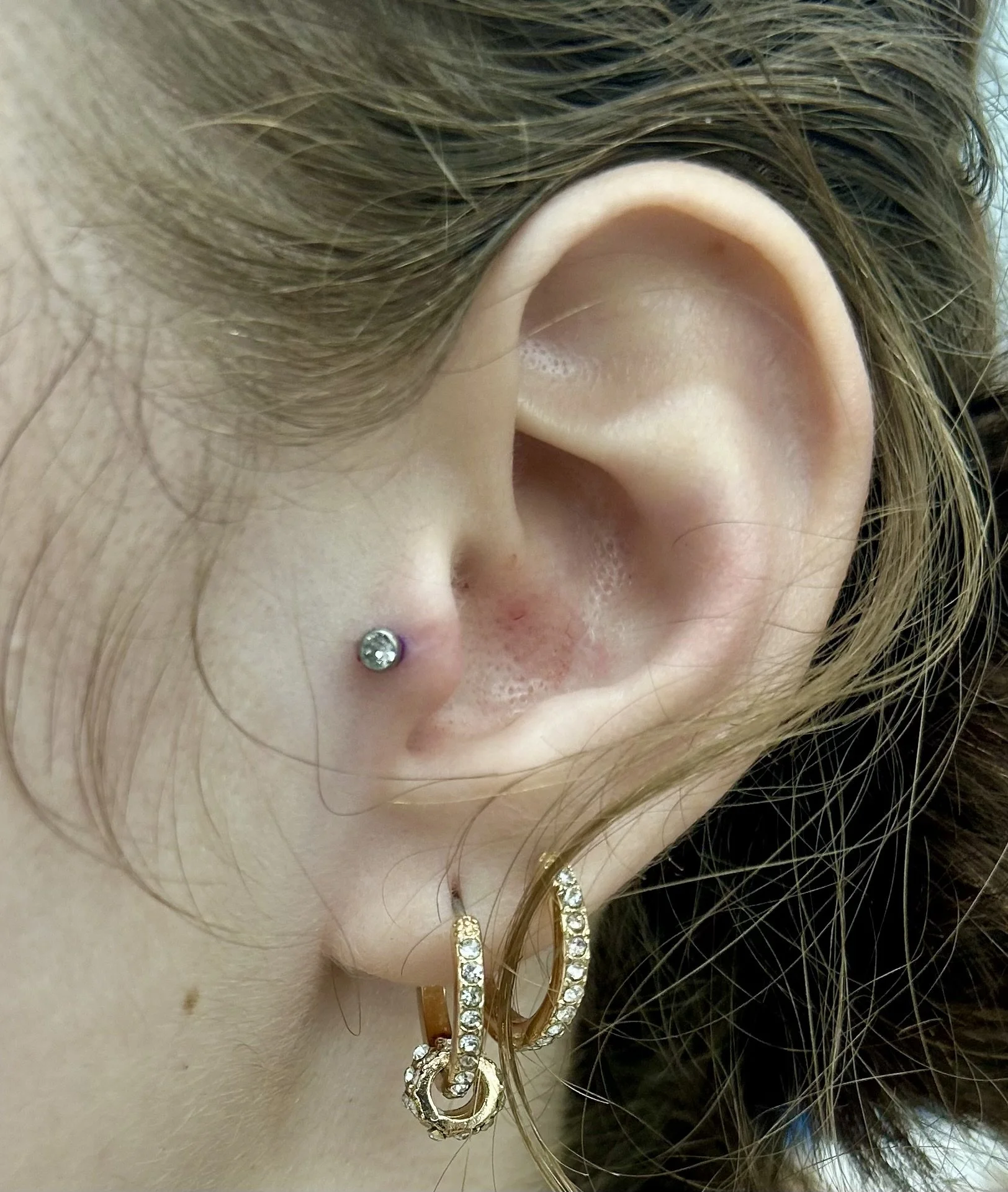 Close-up of a person's ear showing a piercing with a jeweled hoop earring, surrounding hair and necklaces visible.