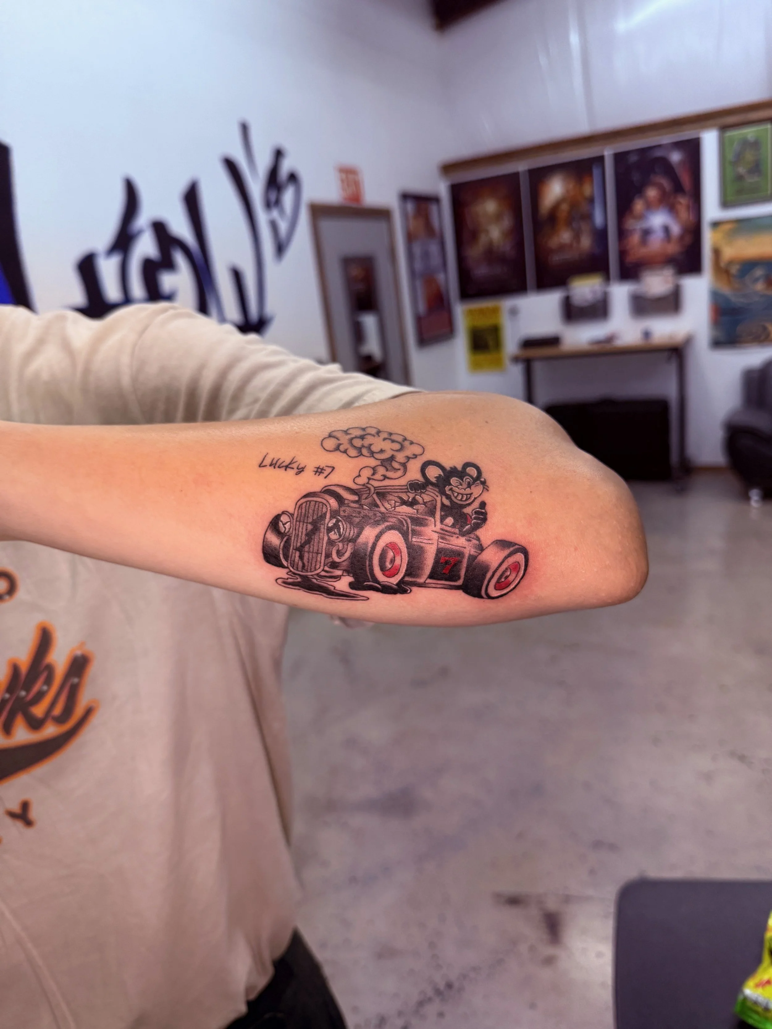 Tattoo of a cartoonish race car with smoke and the text 'Lucky #7' on a person's arm, in an indoor space with artwork on the walls.