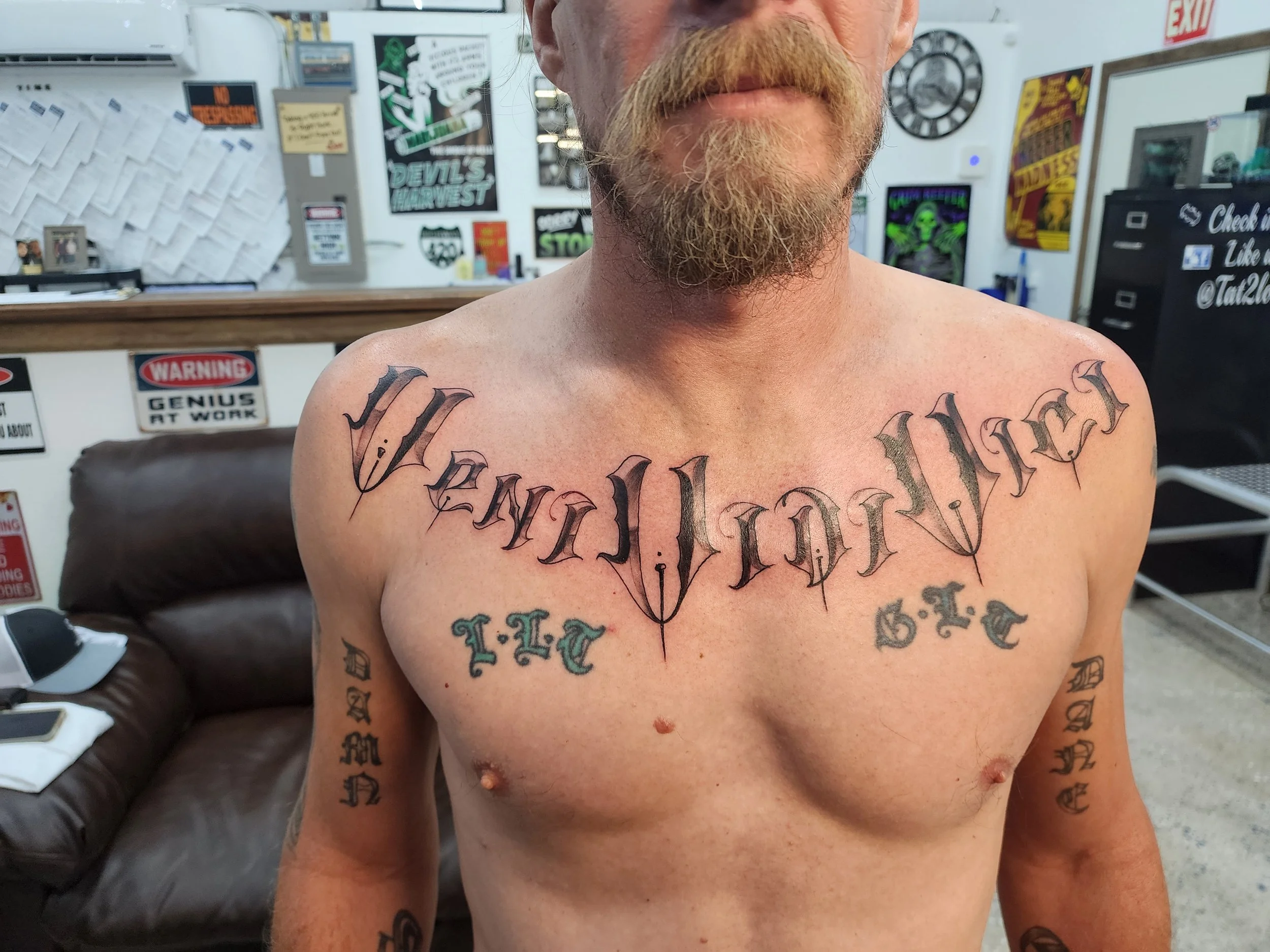 A shirtless man with tattoos on his chest and arms, standing in a tattoo parlor with various signs and posters in the background.