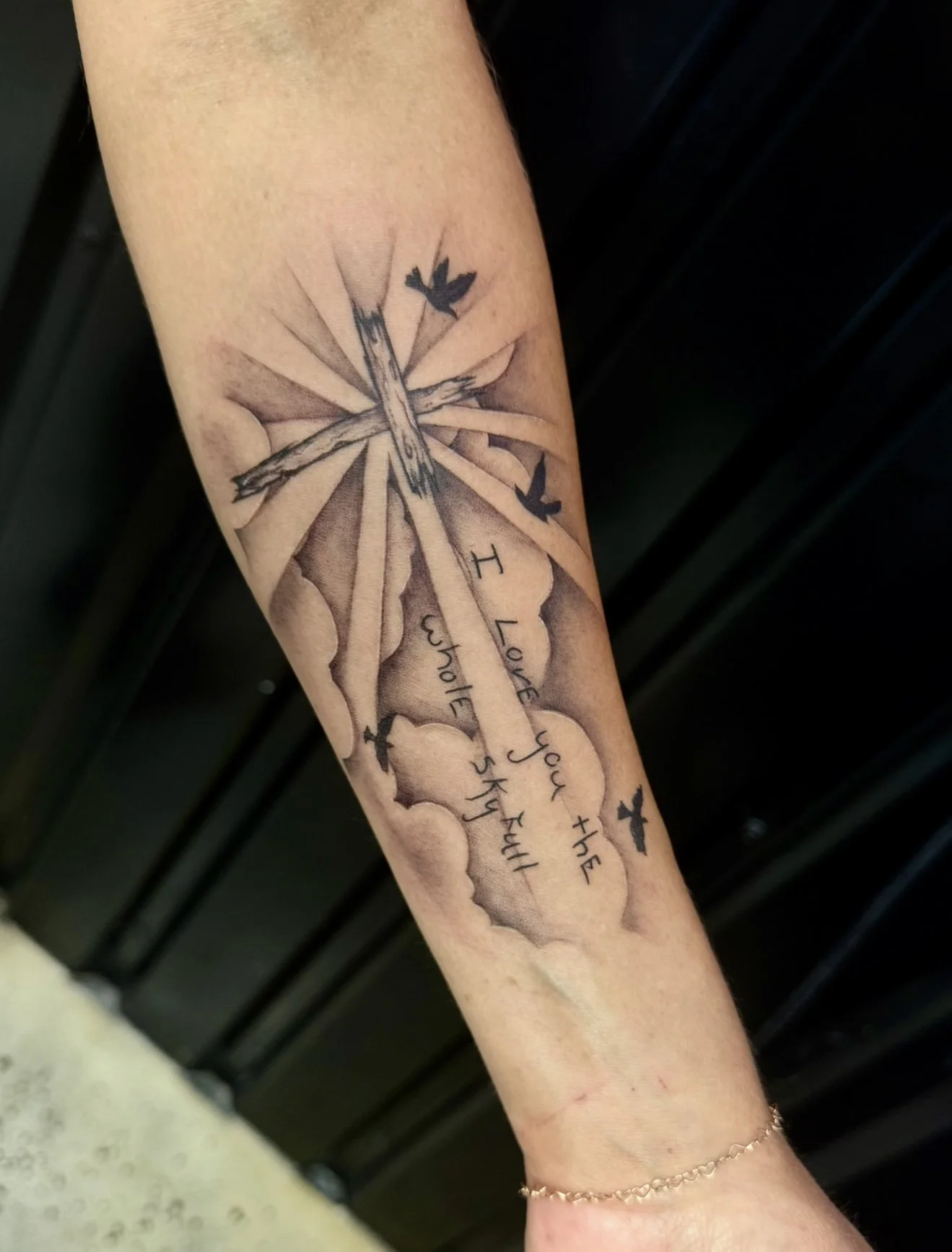 Tattoo of a cross with a gradient background, clouds, and flying birds, with the quote 'I loved you the whole sky full' written underneath.