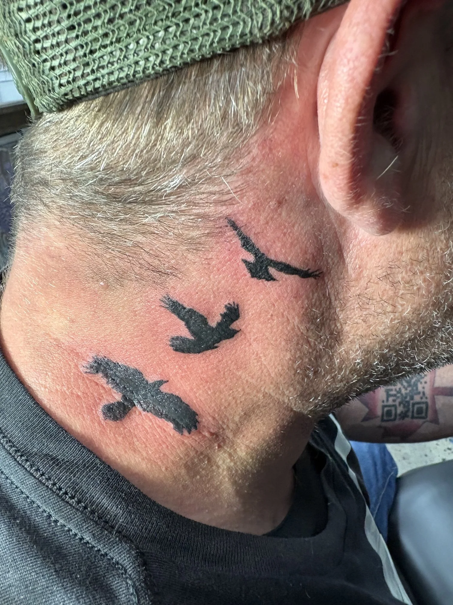 Close-up of a person's side neck and face showing tattoos of black flying birds and some facial hair, with partial view of an ear and a green cap.