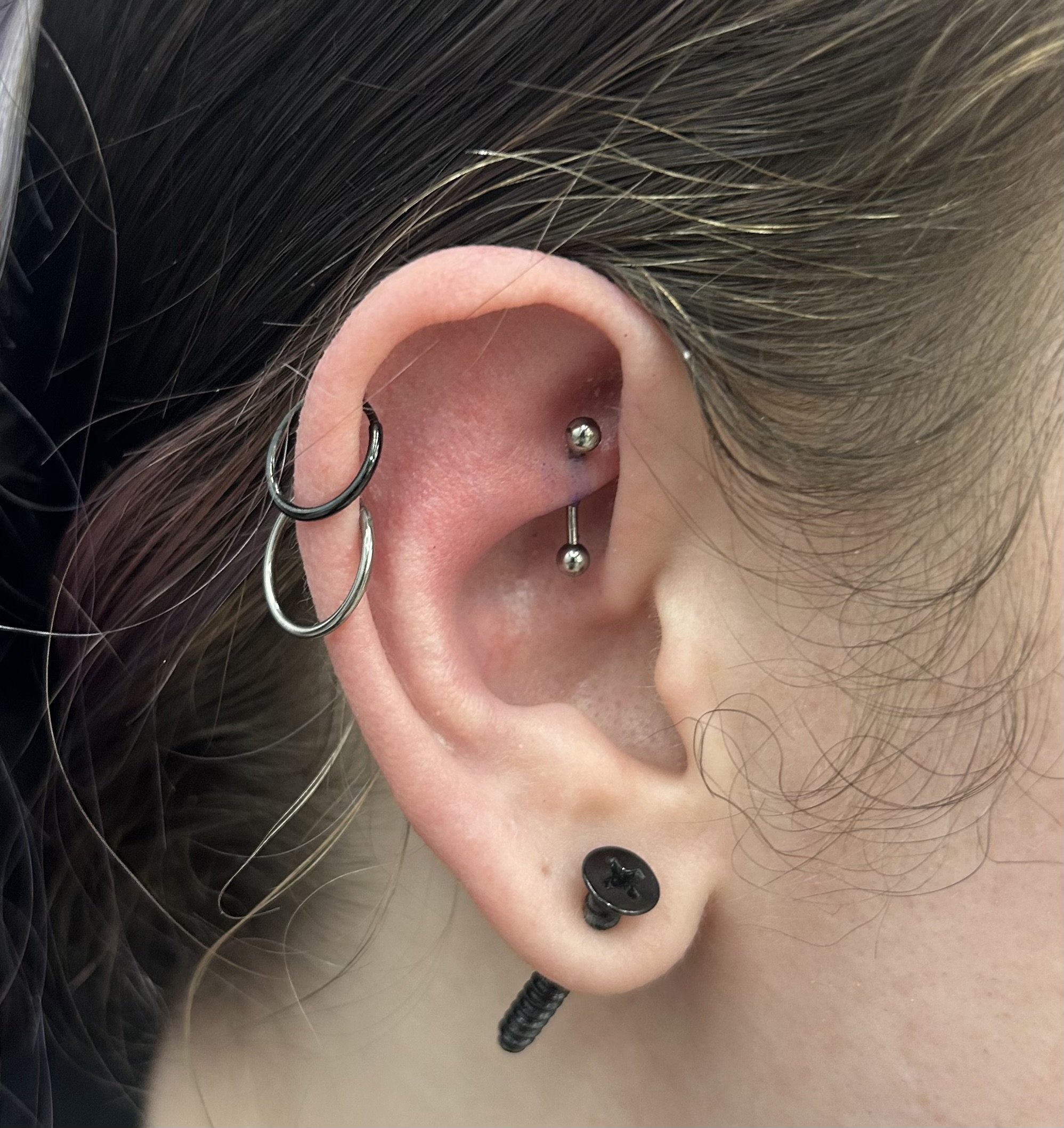 Close-up of a person's ear with multiple piercings, including a small hoop and a ball closure ring, with brown hair and black clothing visible.