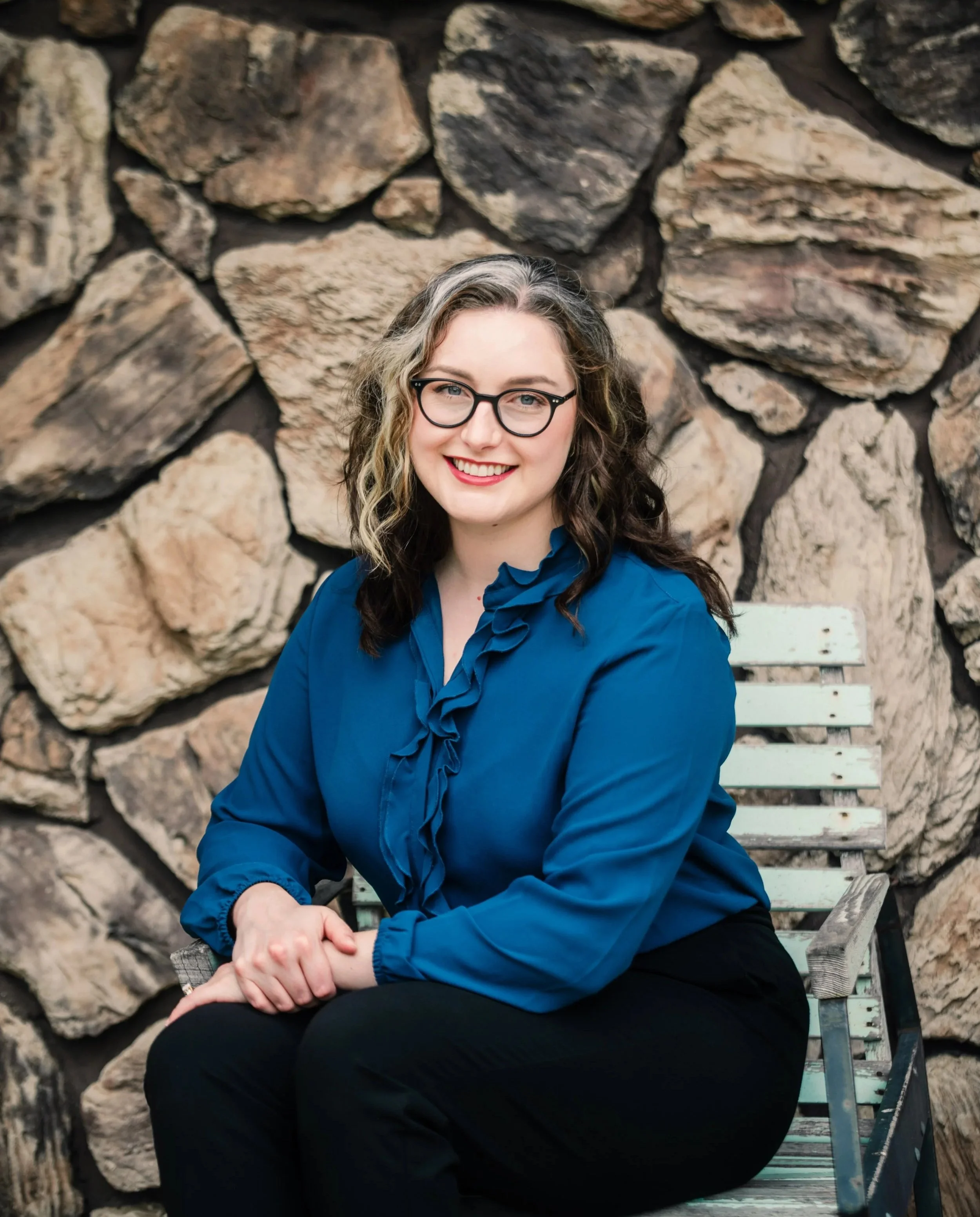 Portrait of Clinical Psychologist Dr. Johanna Wood wearing glasses and a blue blouse seated on a weathered bench against a stone wall