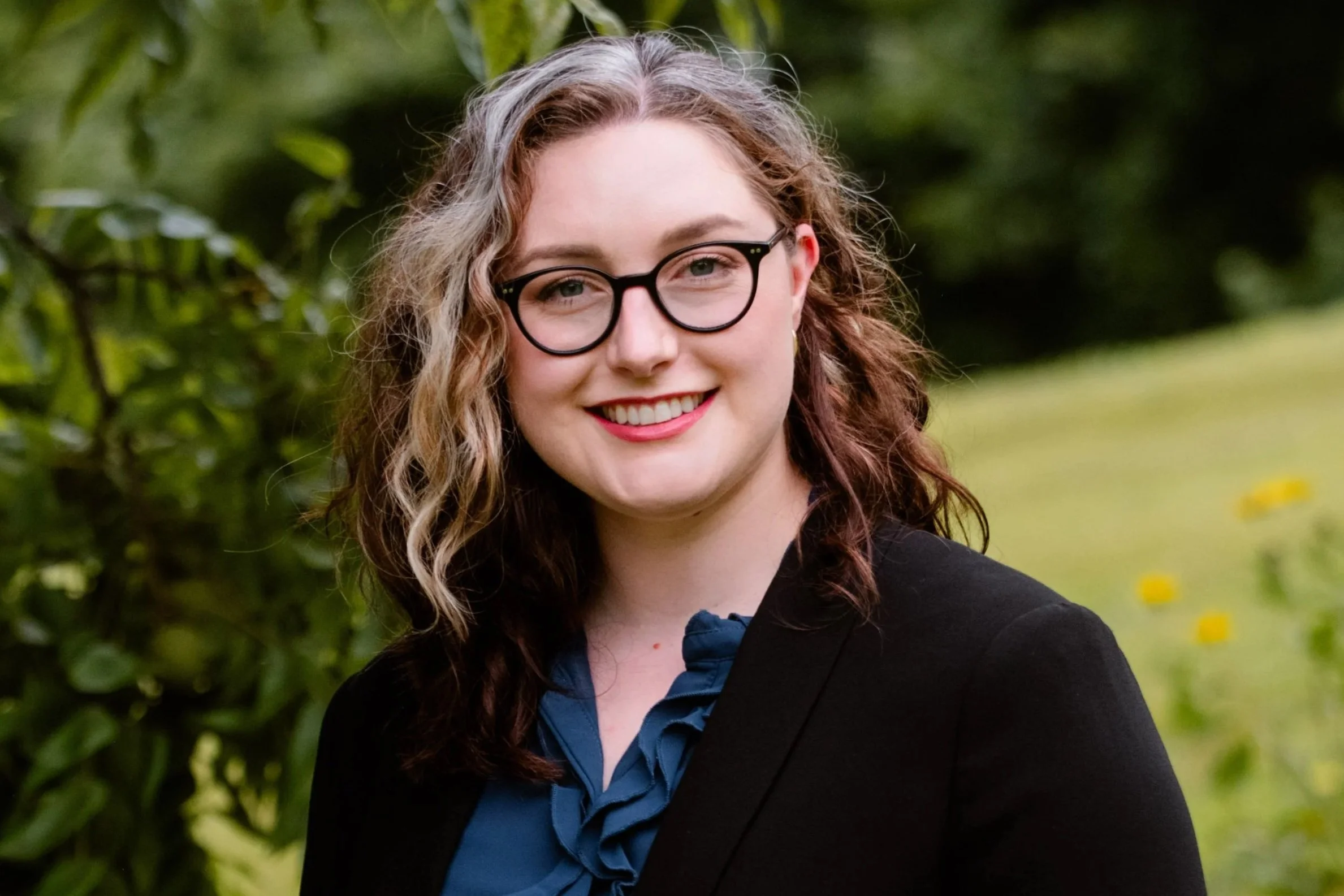 Photo of Dr. Wood, a smiling woman with glasses and curly hair standing outdoors on a grassy area with trees and yellow flowers in the background.