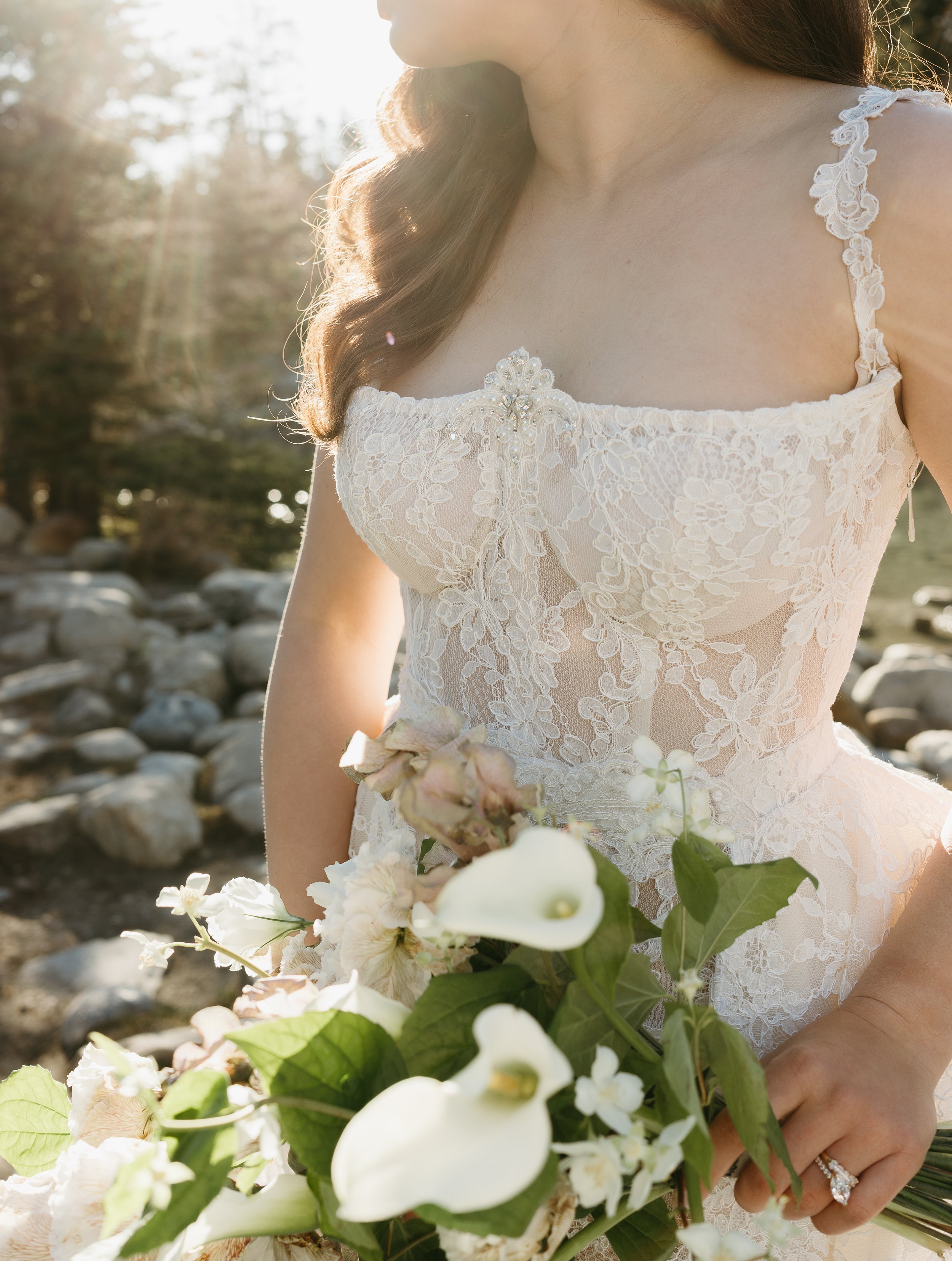 A bride's wedding dress' torso is captured as she holds her bouquet and looks into the distance of a mountain landscape. June 2025 Lake Louise Elopement/Wedding Editorial Session with KC Photography