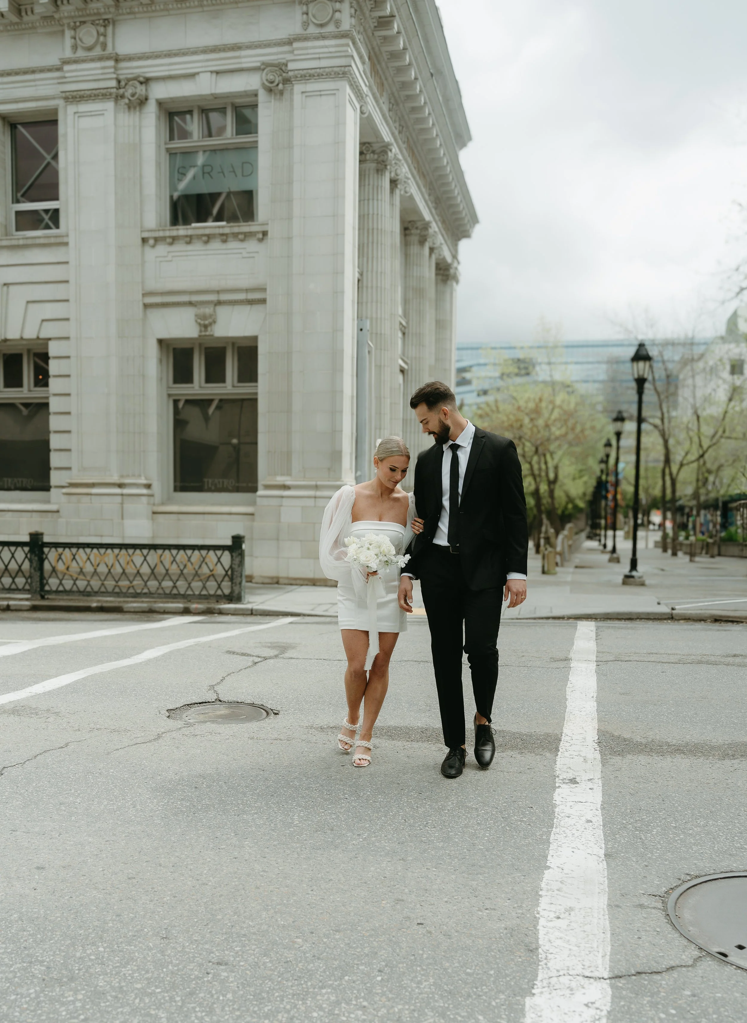 A bride holds her grooms arm as they cross the street of Downtown Calgary. Downtown Calgary Elopement/Wedding Session with KC Photography
