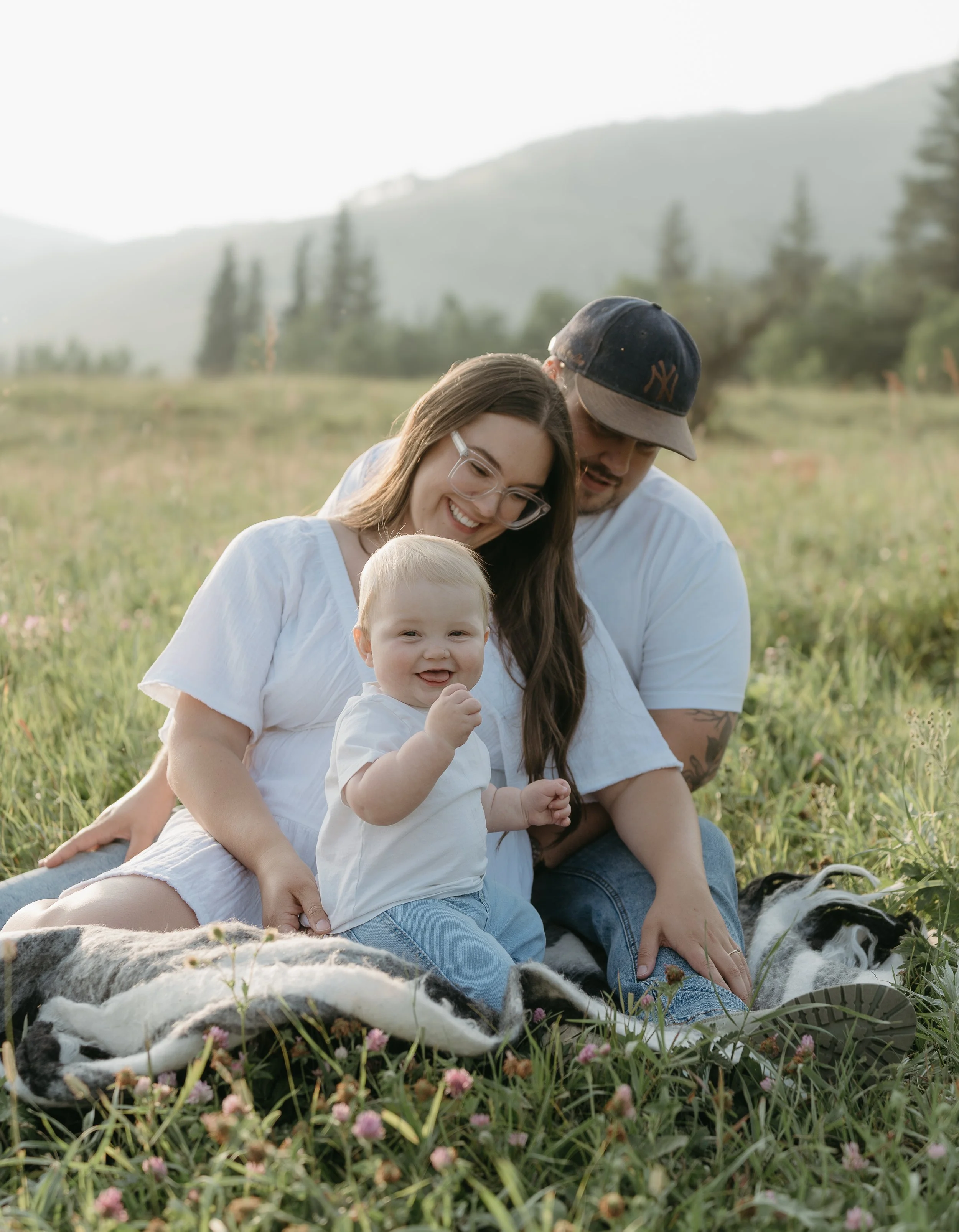 A mother sits in the lap of her husband on a blanket in a field surrounded by mountains while they smile at their infant who sits in front of them, smiling at the camera. Kananaskis Lifestyle/Family Session with KC Photography