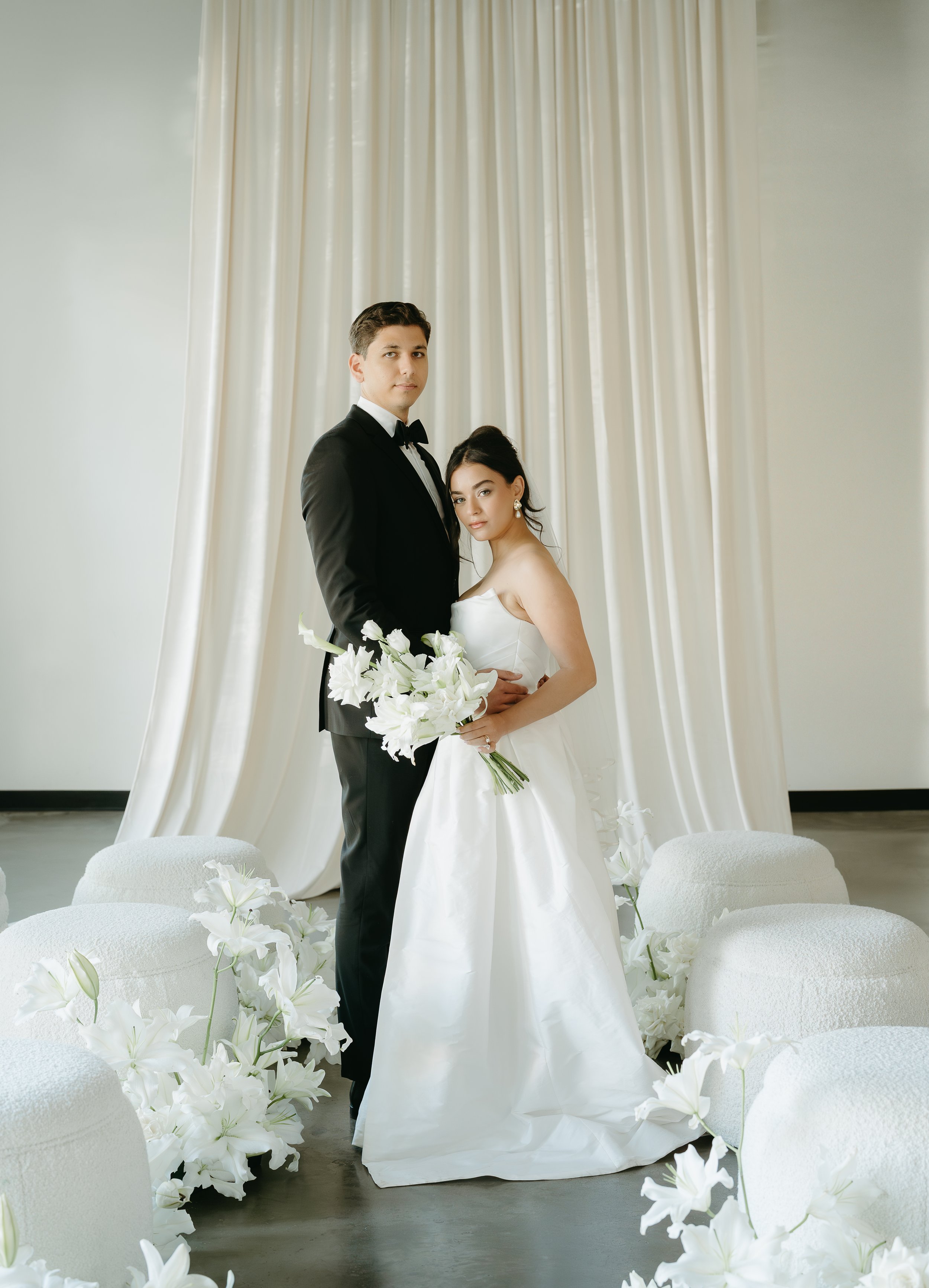 A bride and groom stand against each other looking into the camera in an aisle of chairs and white lily's. July 2025 Calgary Elopement/Wedding Editorial Session with KC Photography