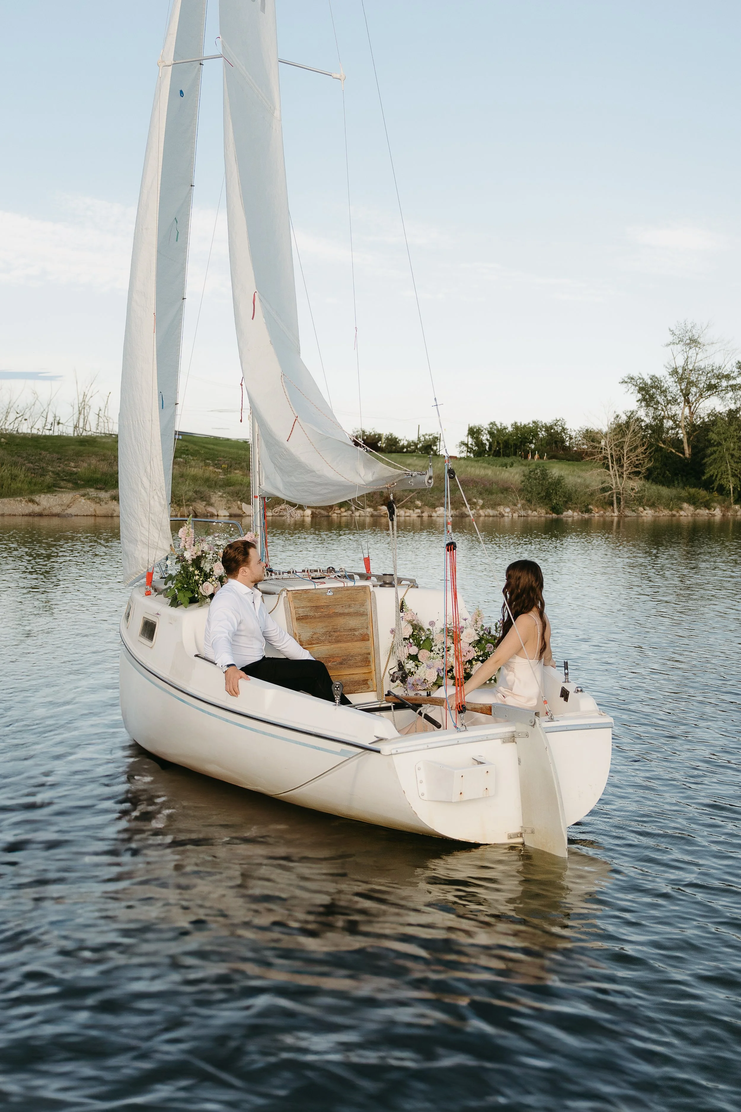 A bride and groom sit on either side of a sailboat with bouquets of flowers floating on the water. August 2025 Elopement/Wedding Sailboat/Sailing Session with KC Photography