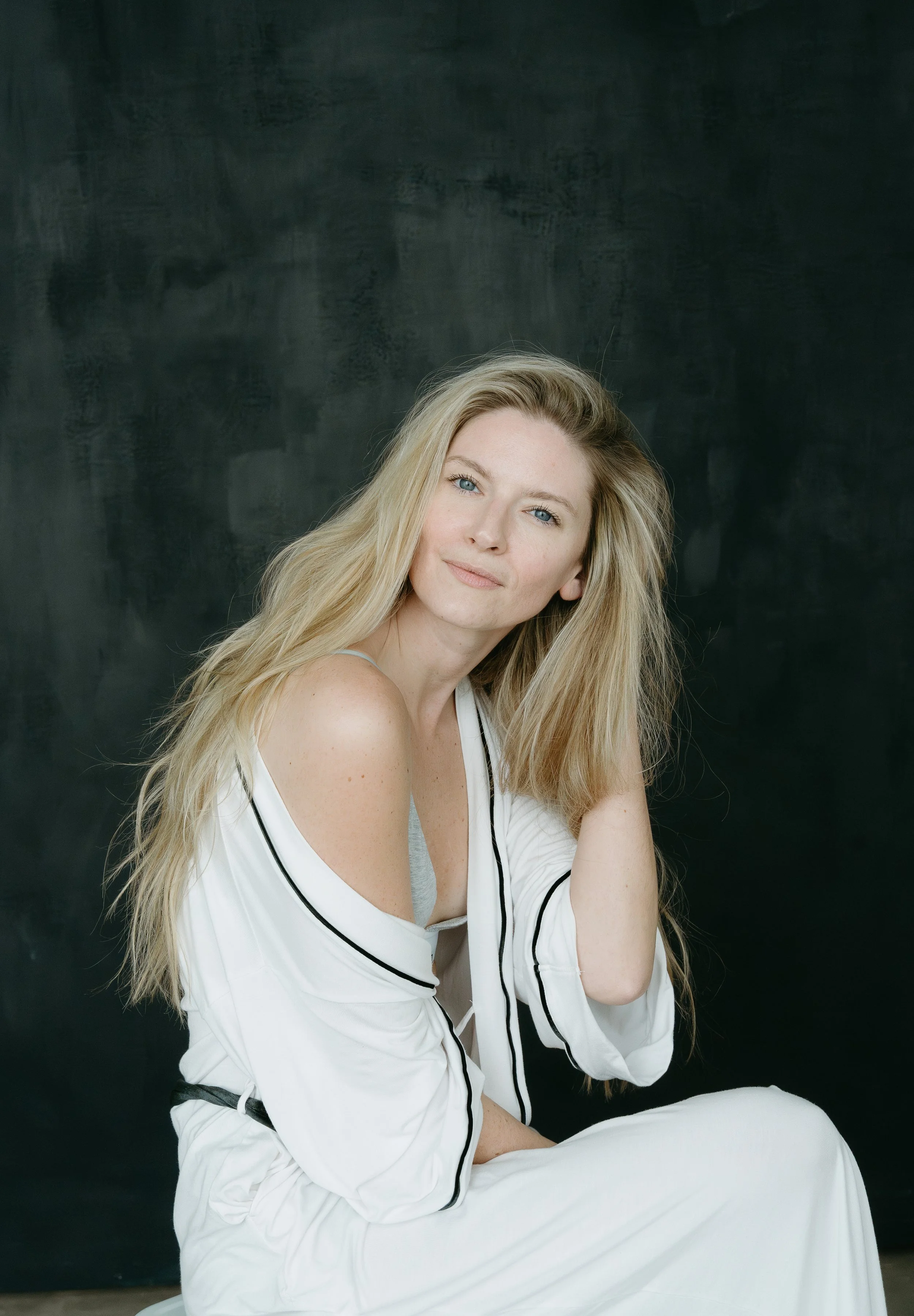A woman sits in front of a dark backdrop and touches her hair as she looks into the camera. Calgary Studio Branding Session with KC Photography