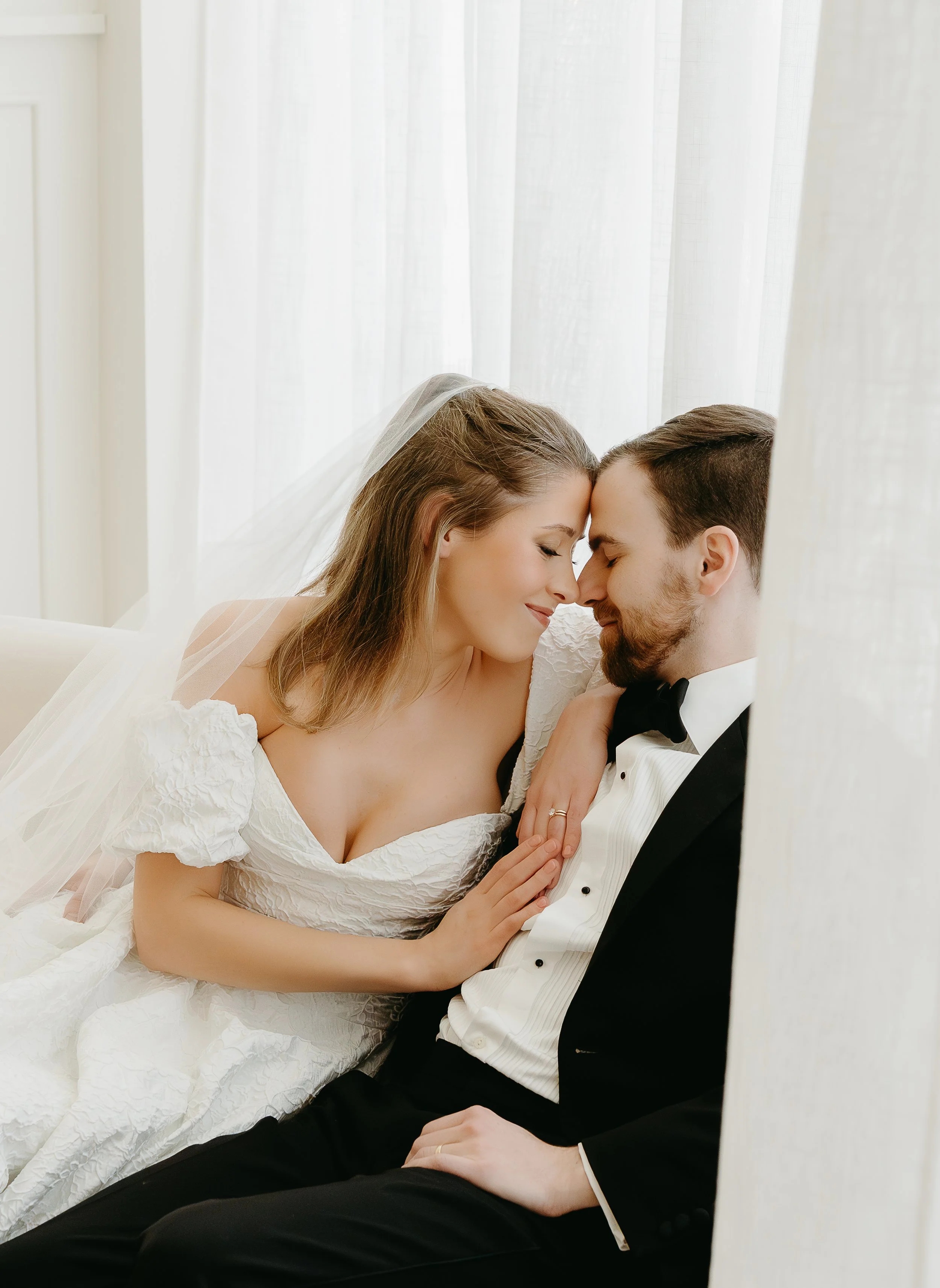 A bride and groom sit on a couch and touch foreheads while smiling. Calgary Elopement/Wedding Editorial Session with KC Photography