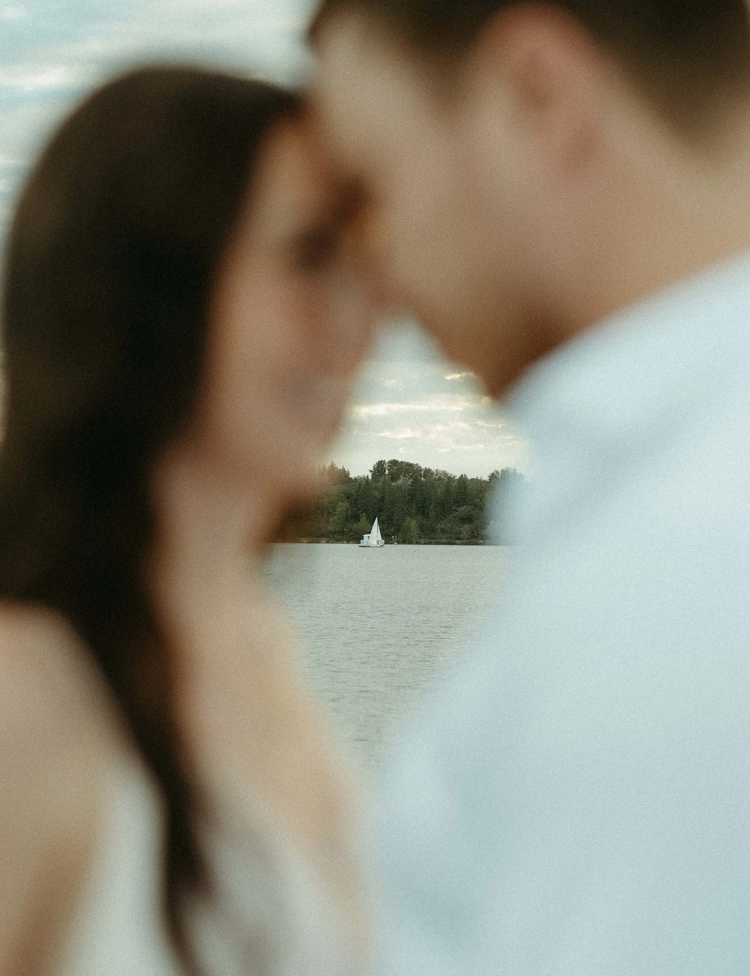 An out of focus bride and groom touch foreheads while an in focus sailboat is spotted on the lake between them. August 2025 Elopement/Wedding Sailboat/Sailing Session with KC Photography