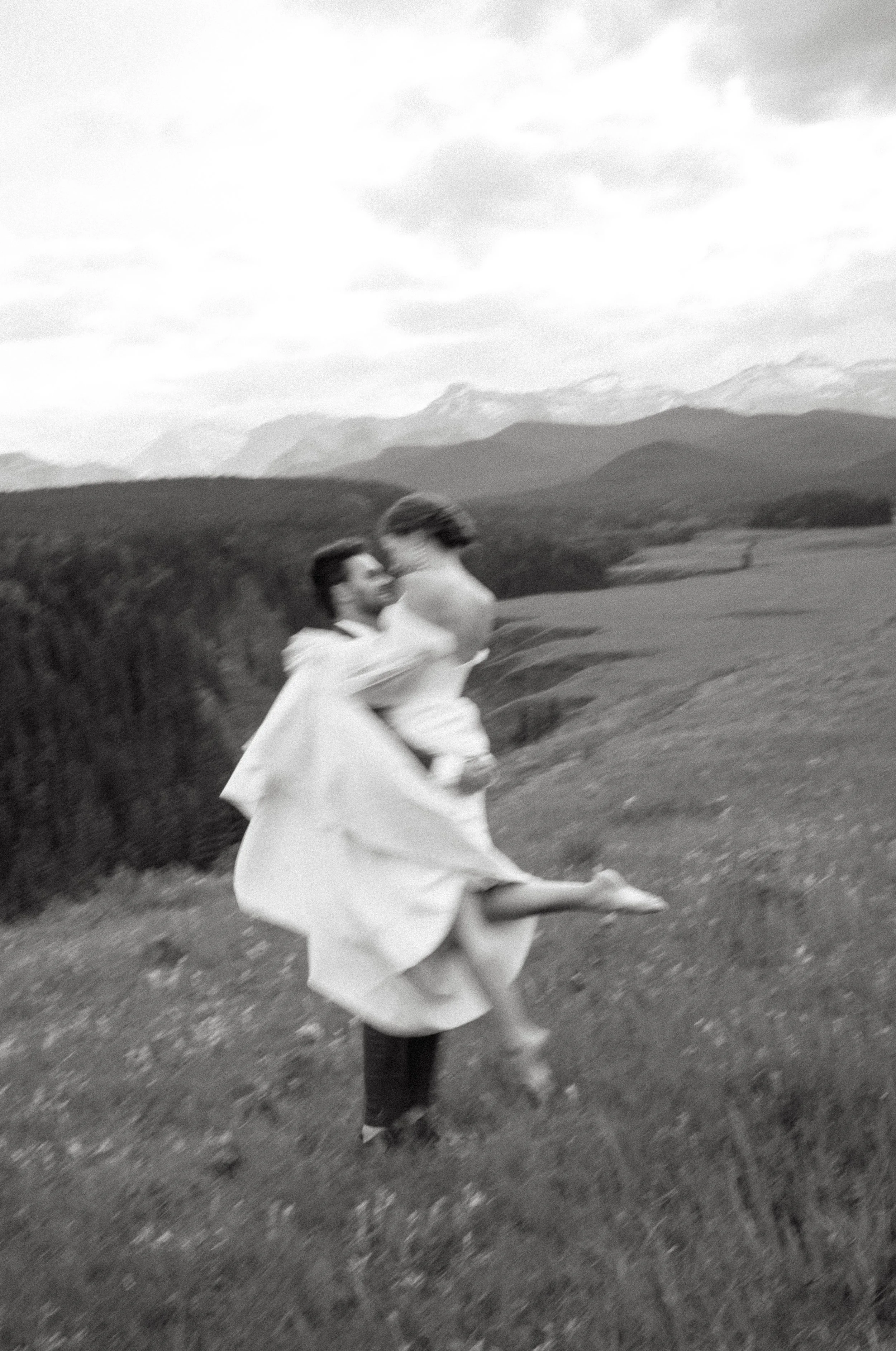 A groom picks up his bride while they look at each other in a field surrounded by mountains. June 2025 Kananaskis Elopement/Wedding Session with KC Photography
