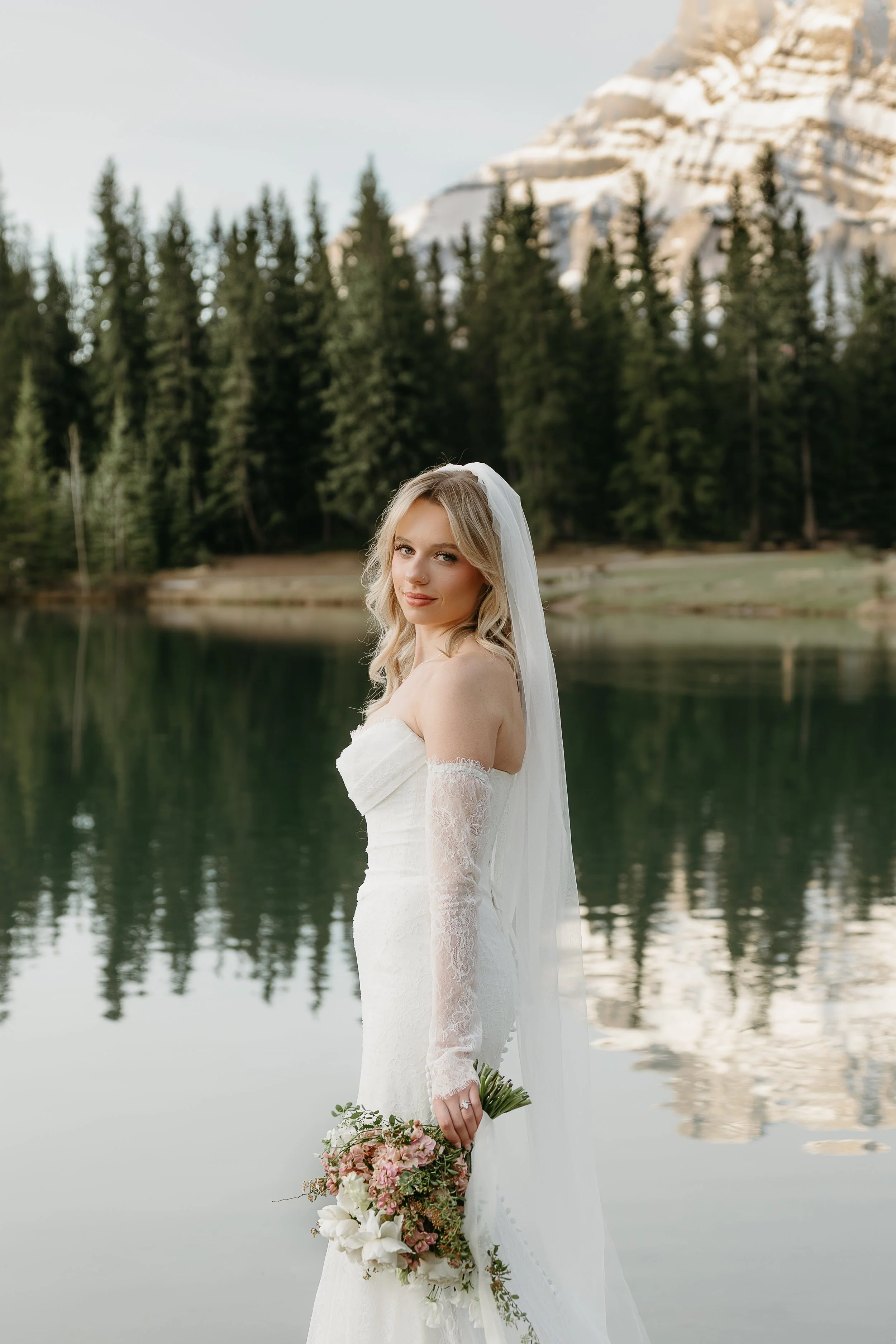 A bride holds a bouquet of flowers and smiles towards the camera beside a lake in Banff surrounded by mountains and pine trees. May 2025 Banff Elopement/Wedding Session with KC Photography