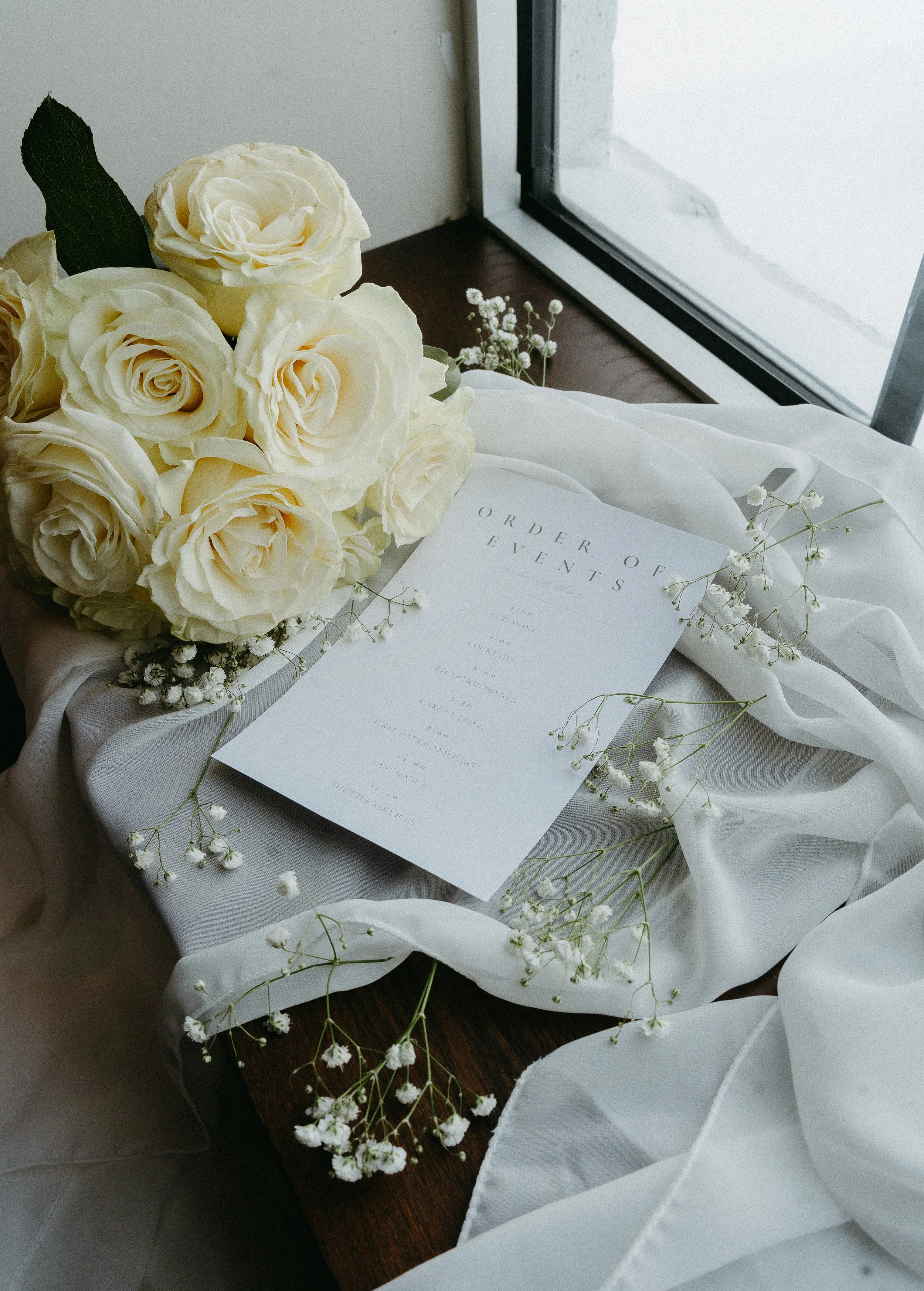 An wedding invitation in surrounded by a bouquet of white roses and scatters baby's breath flowers on a window sill. Calgary Elopement/Wedding Session with KC Photography