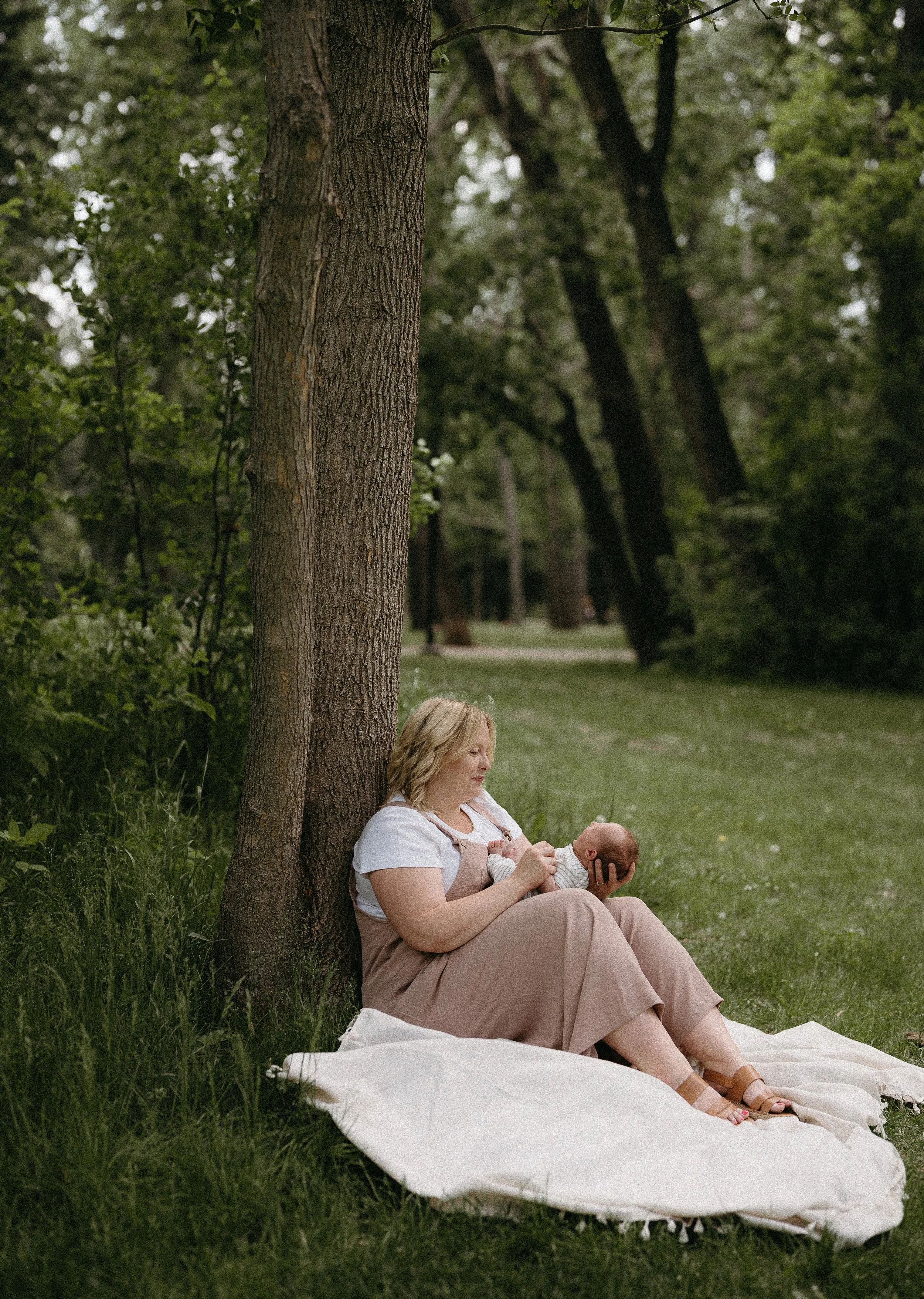 A women sits against a tree in a field as she holds and looks down at her baby. Calgary River Lifestyle/Family/Newborn Session with KC Photography
