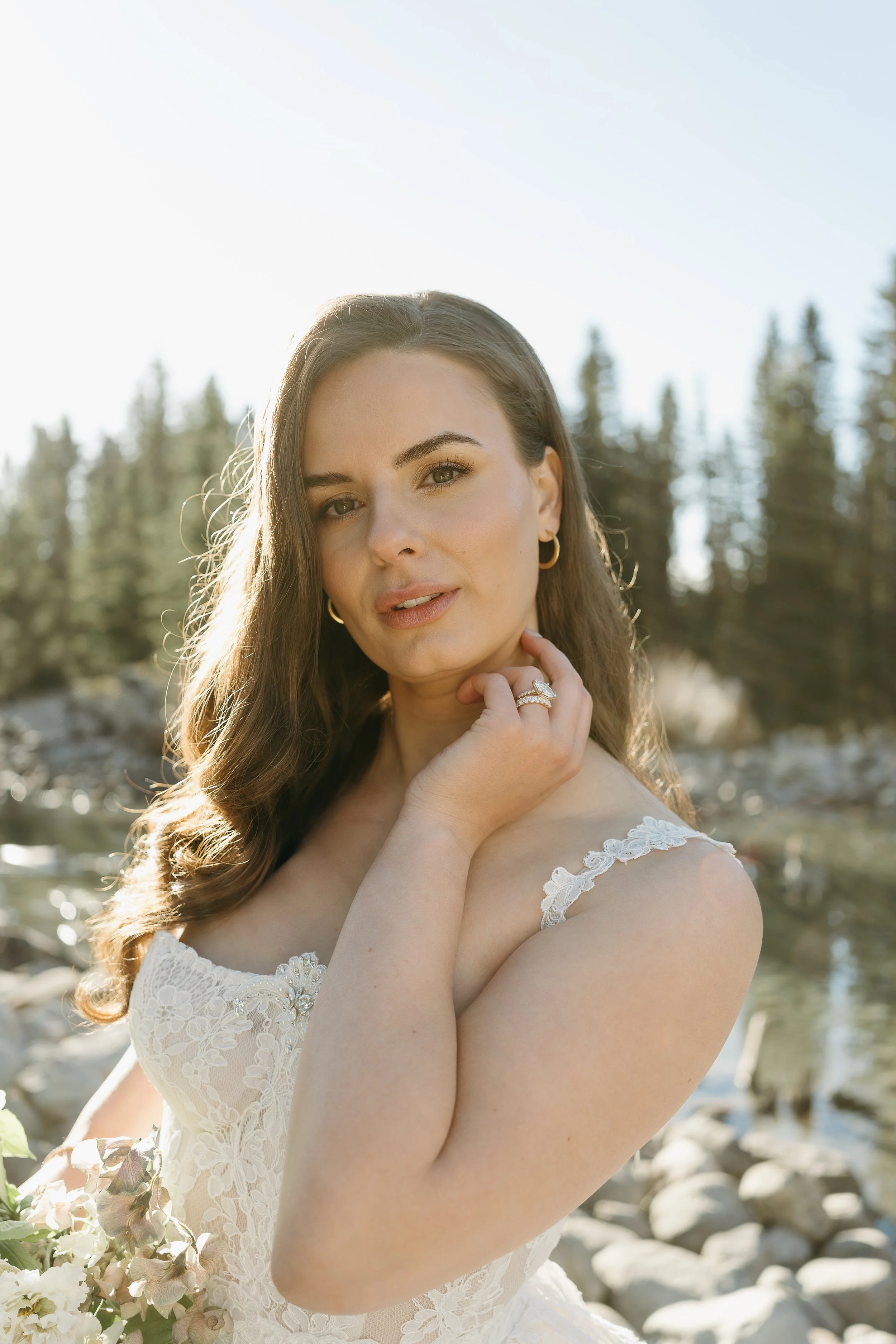A bride touches her neck as she look into the camera on the edge of a mountain creek. June 2025 Lake Louise Elopement/Wedding Editorial Session with KC Photography