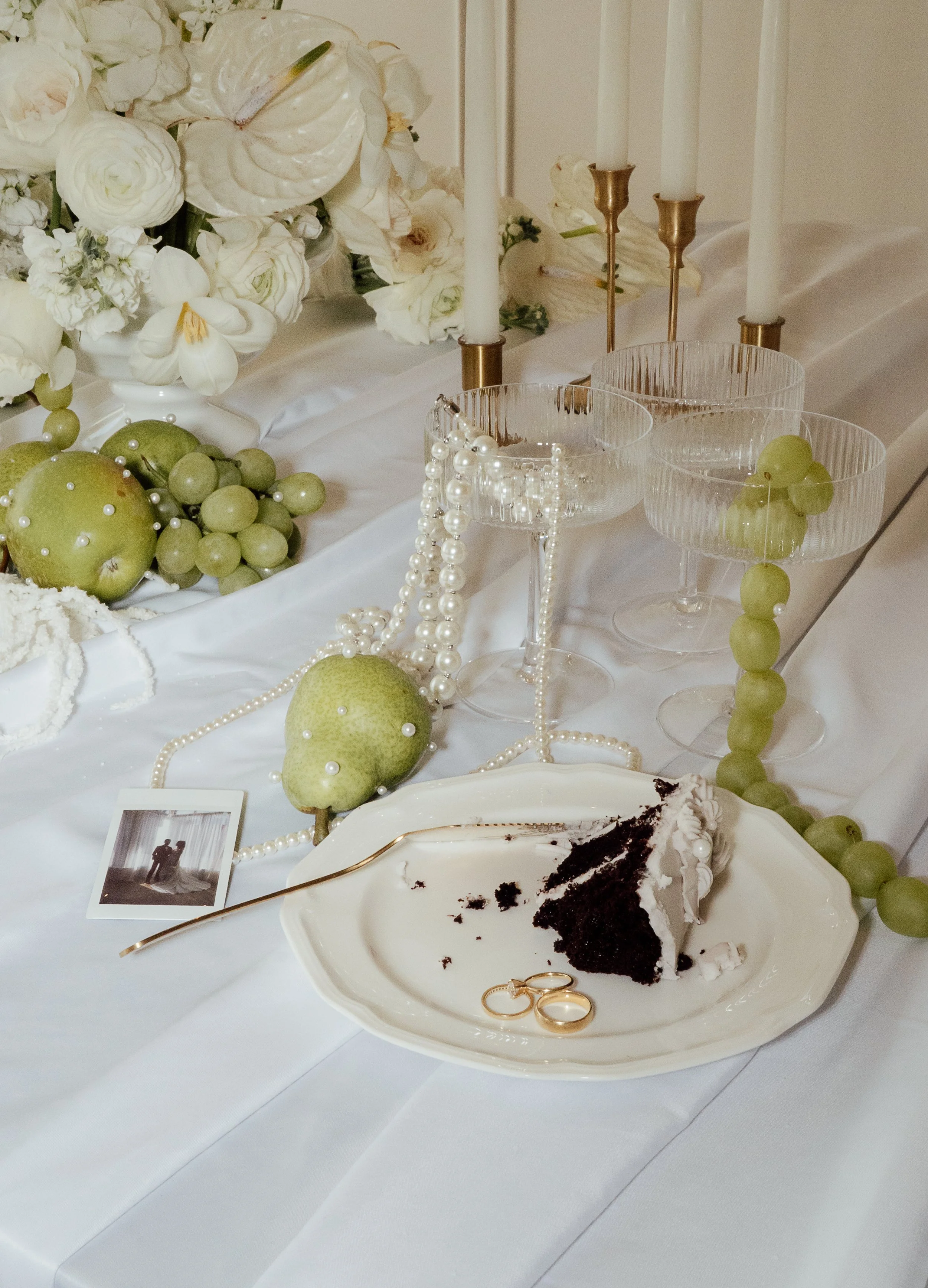 A half eaten piece of wedding cake on a plate with wedding rings is placed on a reception table covered with flowers, champagne glasses, green fruits, and pearls. Calgary Elopement/Wedding Editorial Session with KC Photography