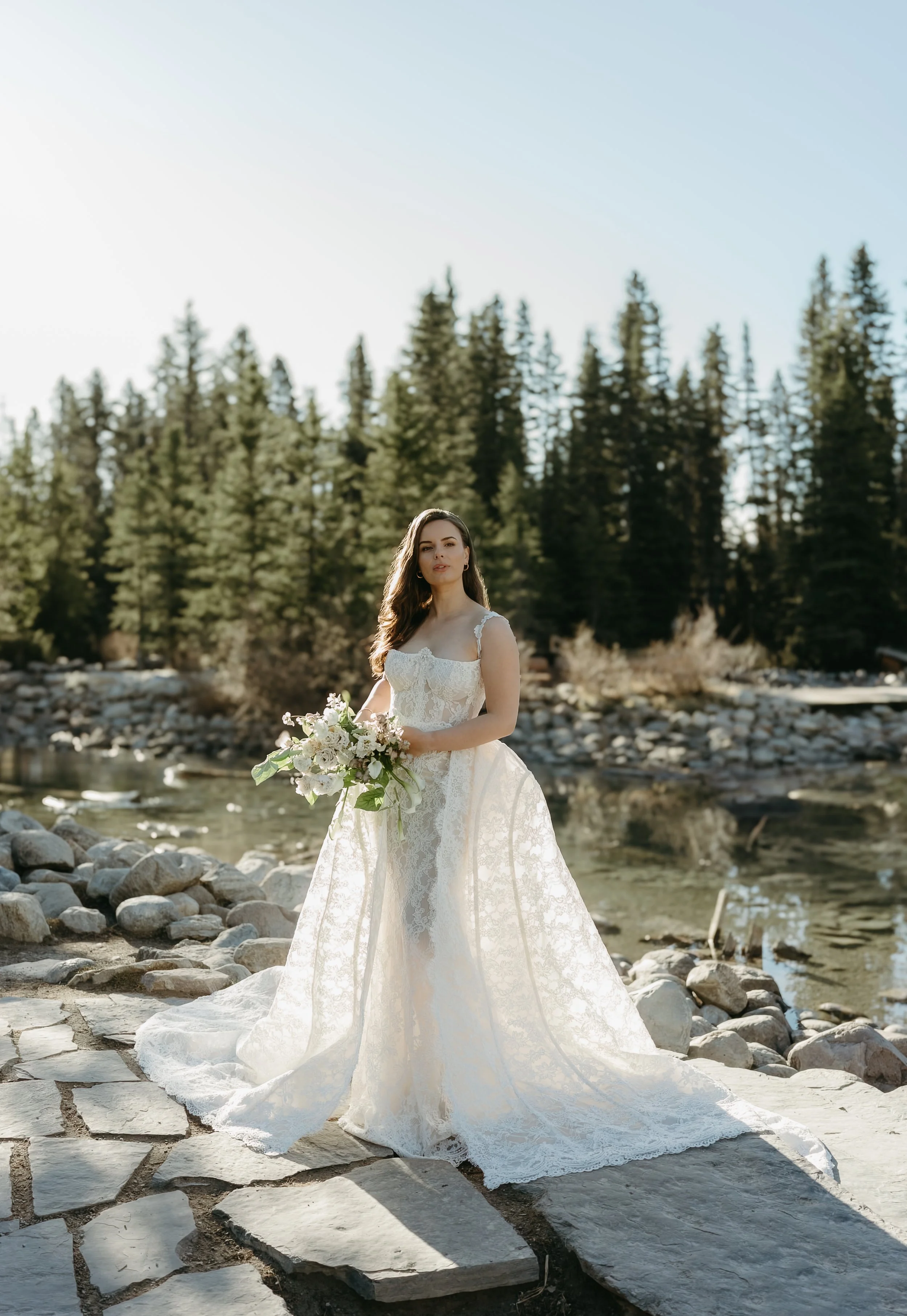 A bride stands on the edge of a mountainous creek holding a bouquet of flowers. June 2025 Lake Louise Elopement/Wedding Editorial Session with KC Photography