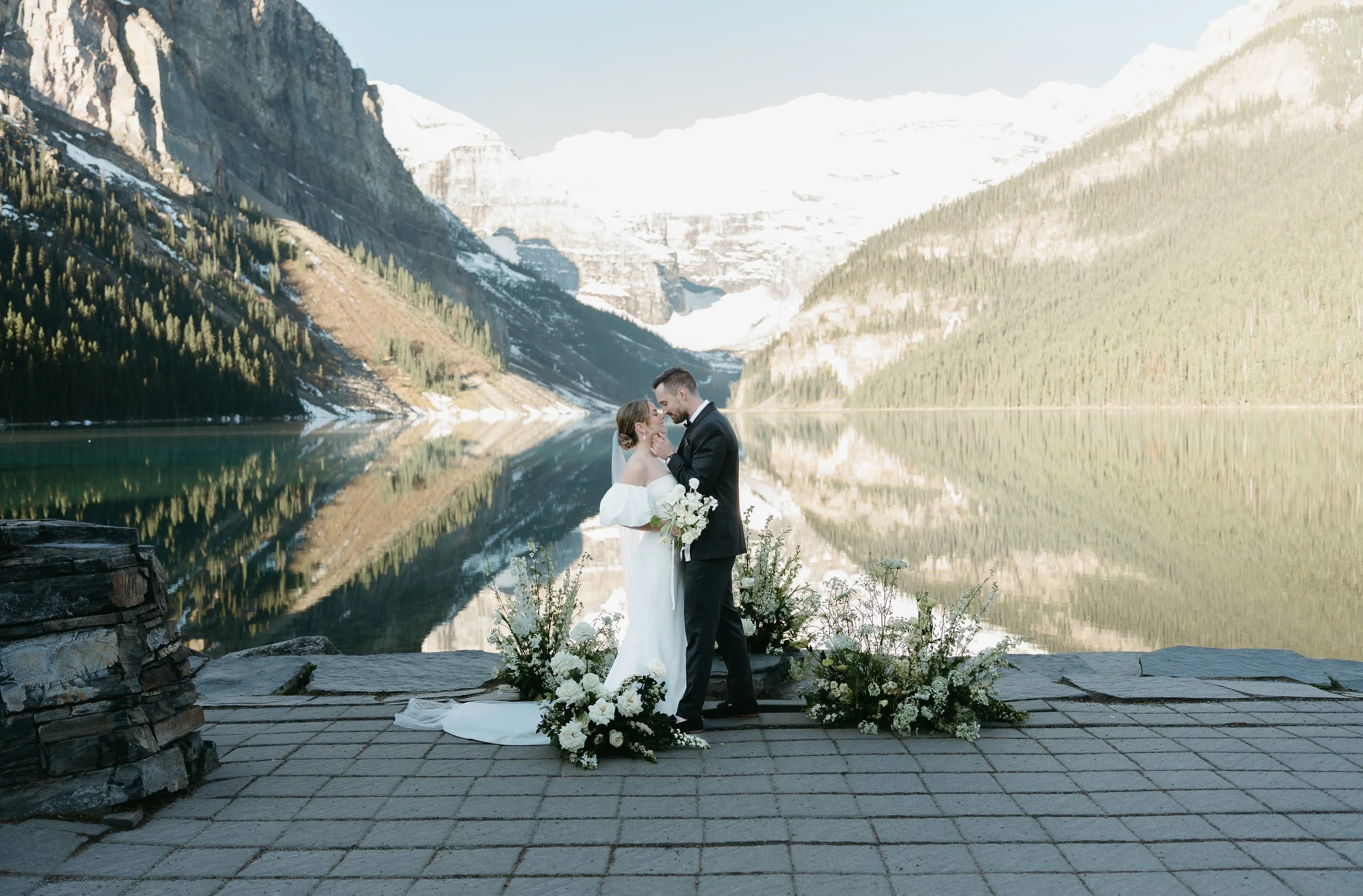 A groom touches the cheek of his bride as he approaches her for a kiss on the edge of a lake surrounded by mountains. August 2025 Canmore/Banff Elopement/Wedding Editorial Session with KC Photography