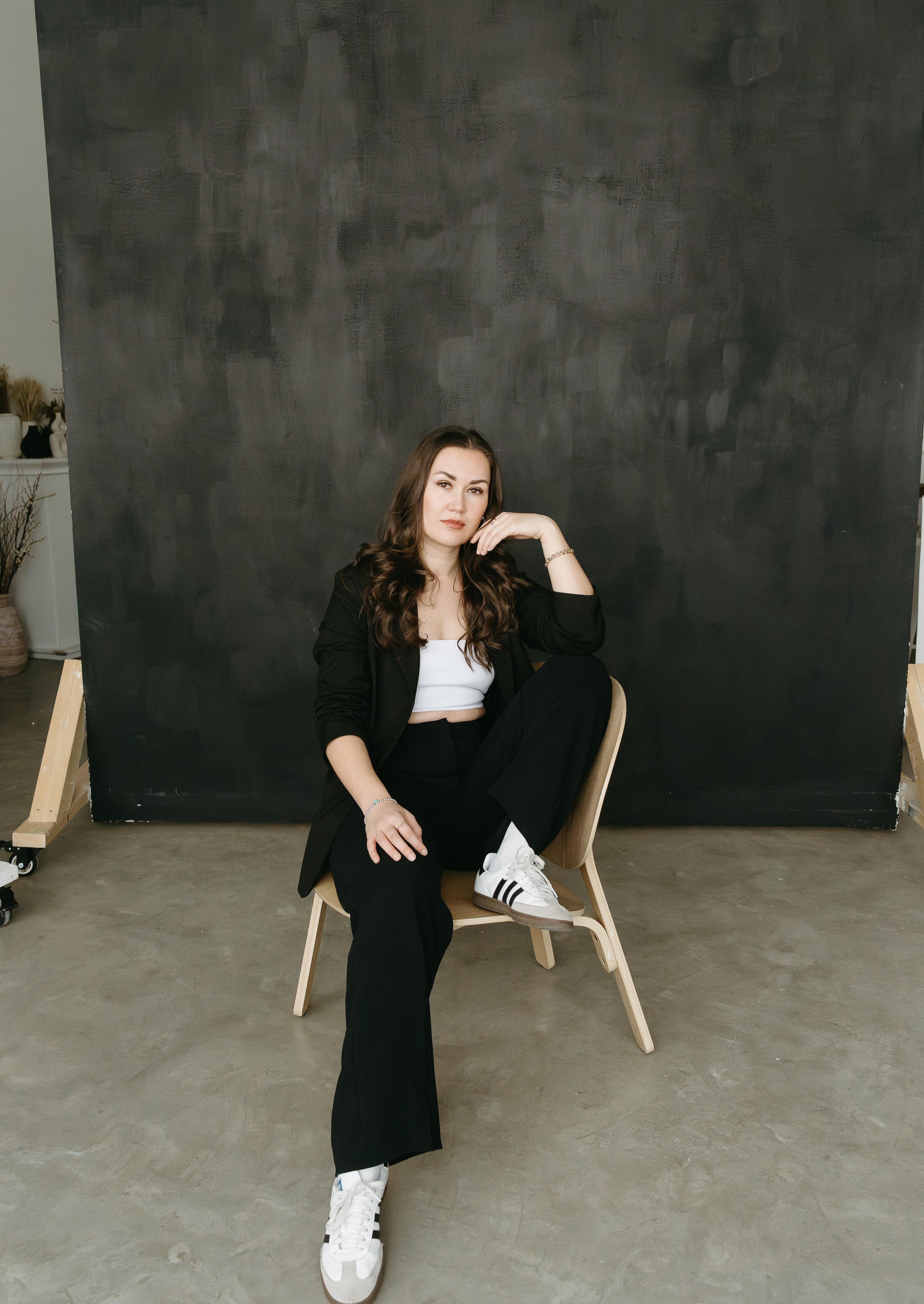 A woman sits in a chair against a dark studio backdrop, with one leg up and resting her elbow on her knee while touching her chin and looking into the camera. Calgary Branding Studio Session with KC Photography