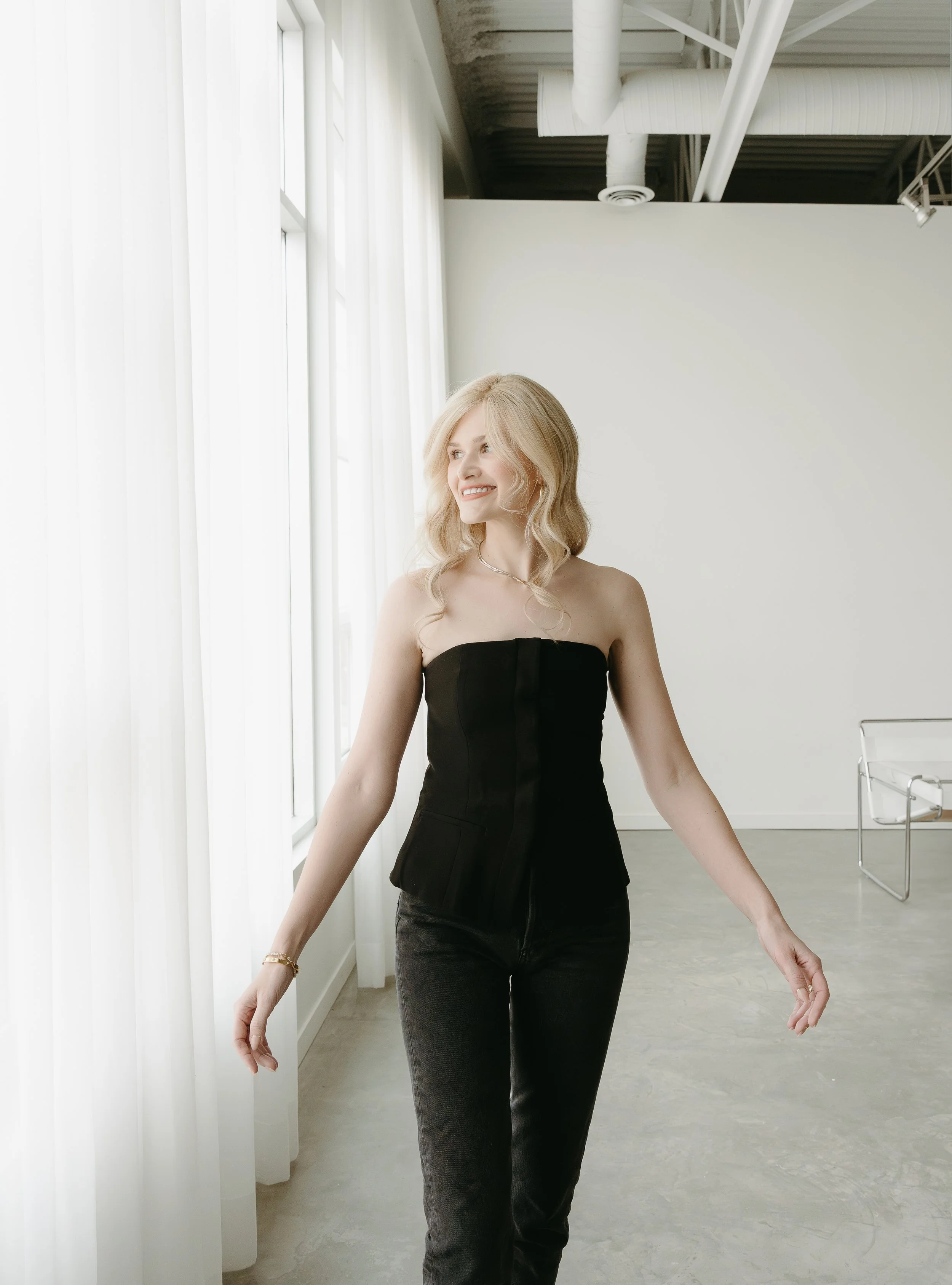 A woman walks along a window of floor length curtains, while holding her arms out beside her, and looking out smiling.Calgary Branding Studio Session with KC Photography