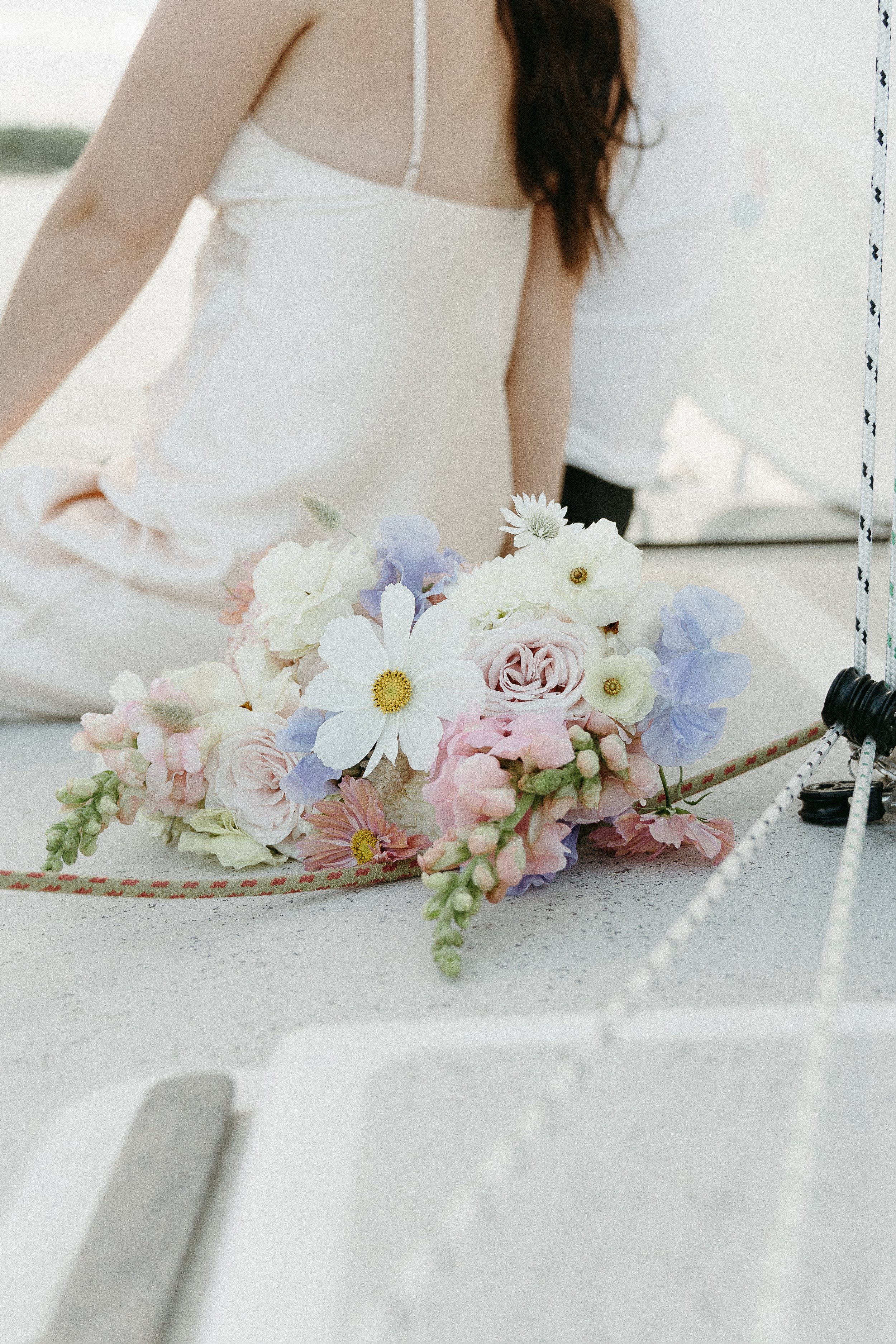 A bouquet of flowers on the back of a sailboat is captured while a couple is spotted sitting beside each other in the background. August 2025 Elopement/Wedding Sailboat/Sailing Session with KC Photography