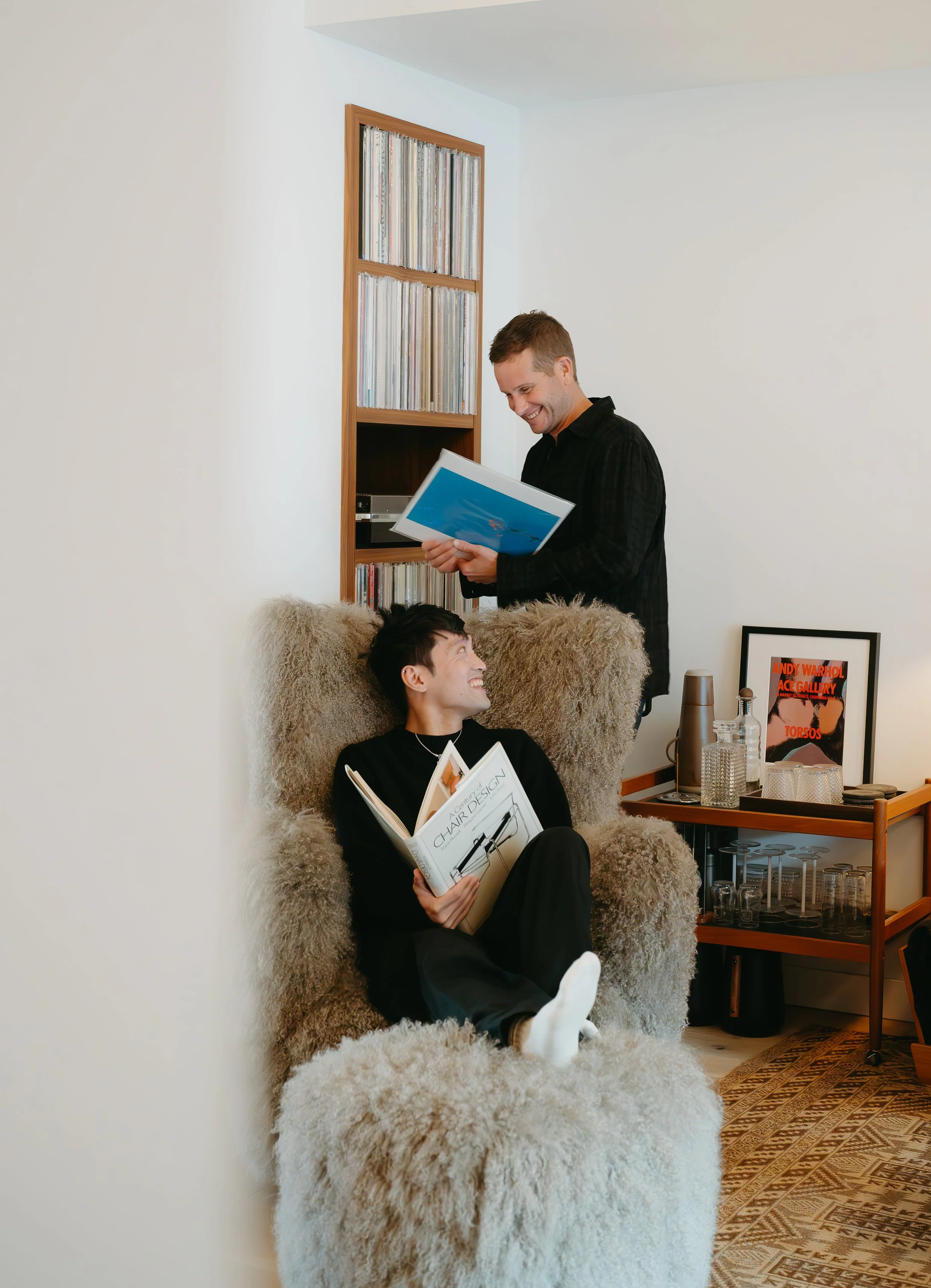 A man sits in a chair holding a book as he looks at his partner standing behind him while they both exchange a smile. July 2025 Lifestyle/Family/Couple Session with KC Photography