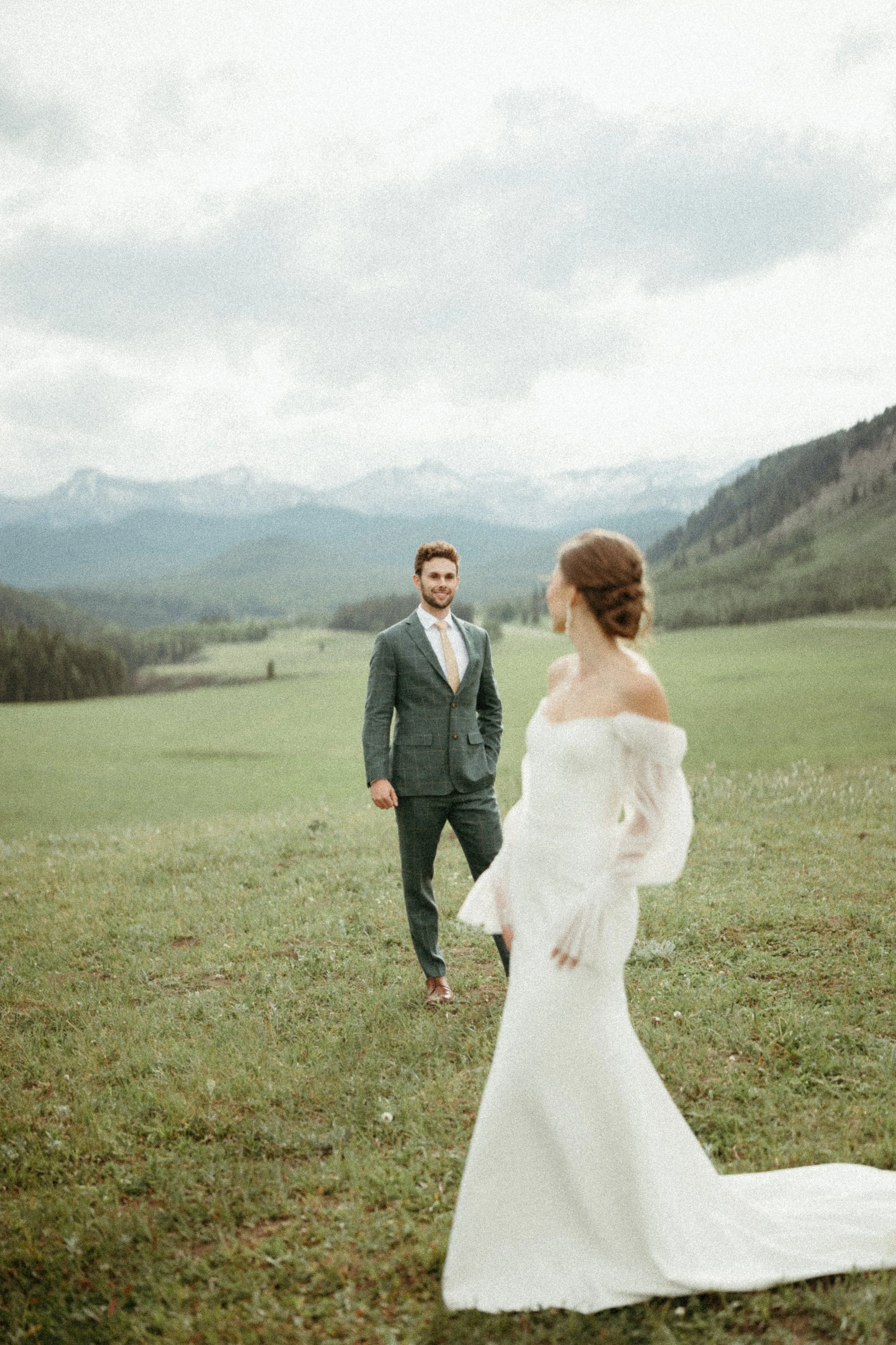 A bride is turned away from the camera, looking back at her husband in a field surrounded by mountains. June 2025 Kananaskis Elopement/Wedding Session with KC Photography
