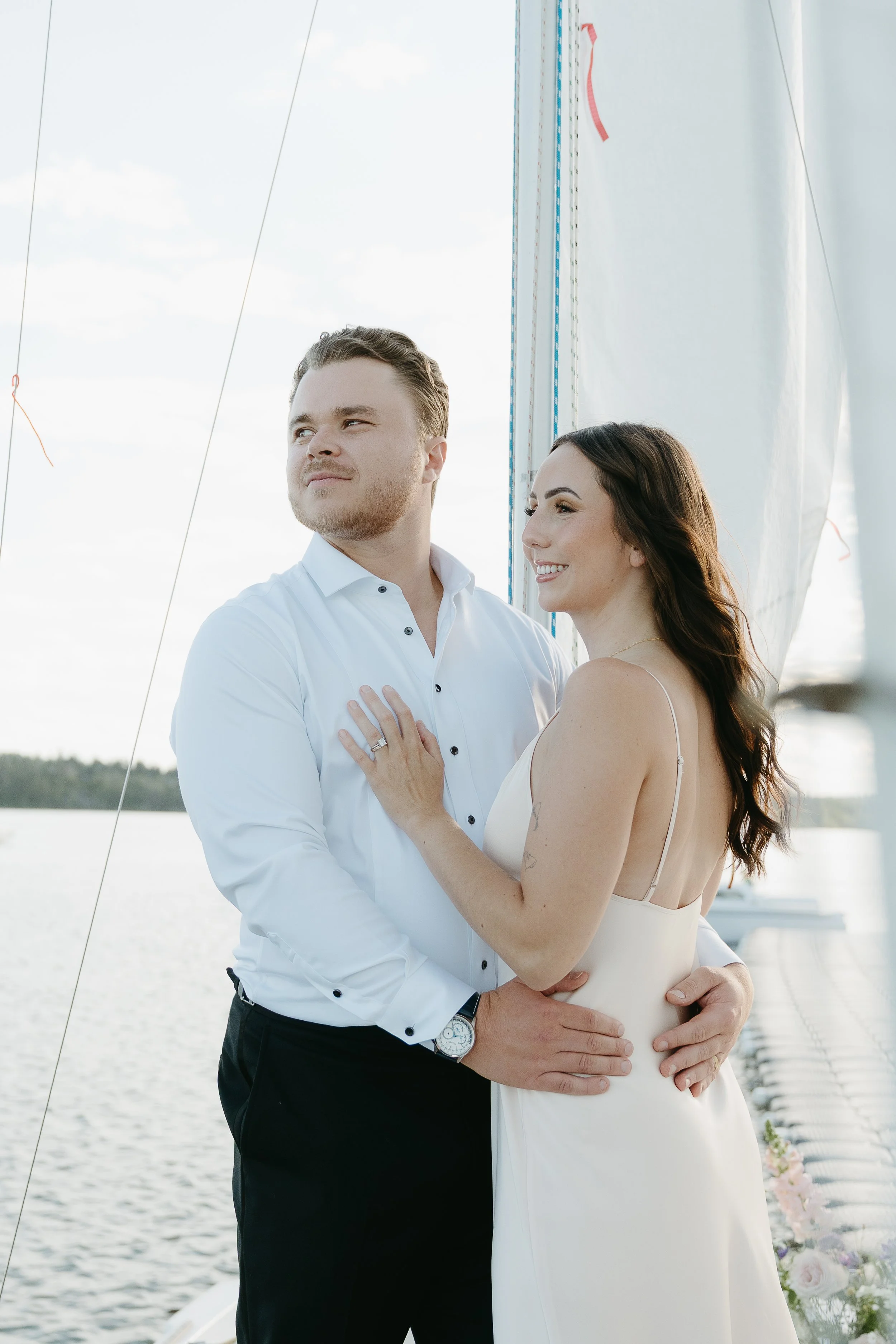 A groom holds his bride in front of the sailing post of a sailboat as they look off into the distance. August 2025 Elopement/Wedding Sailboat/Sailing Session with KC Photography
