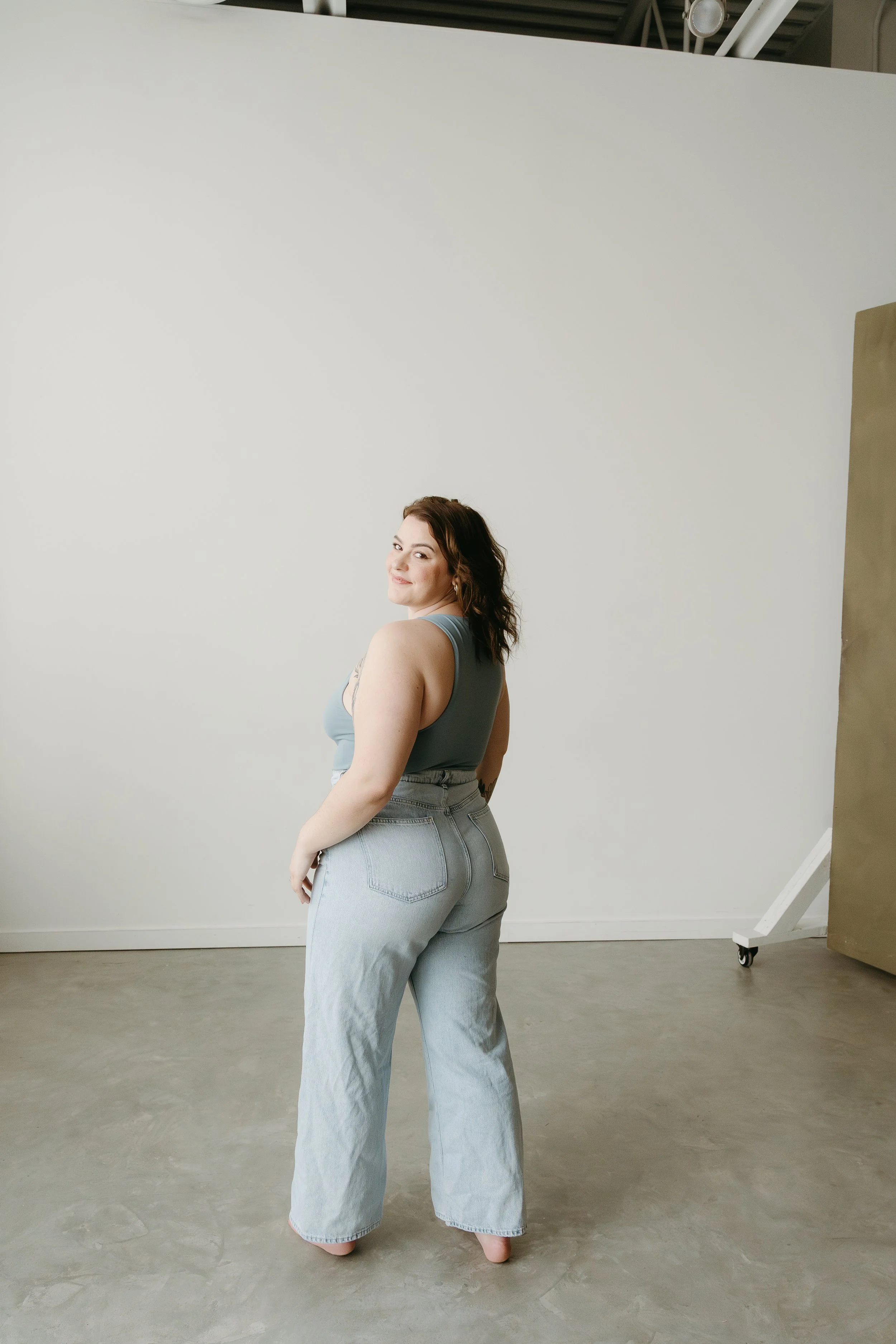 A woman stands with her back towards the camera and smiles over her shoulder at the camera in a studio setting. Calgary Branding Studio Session with KC Photography