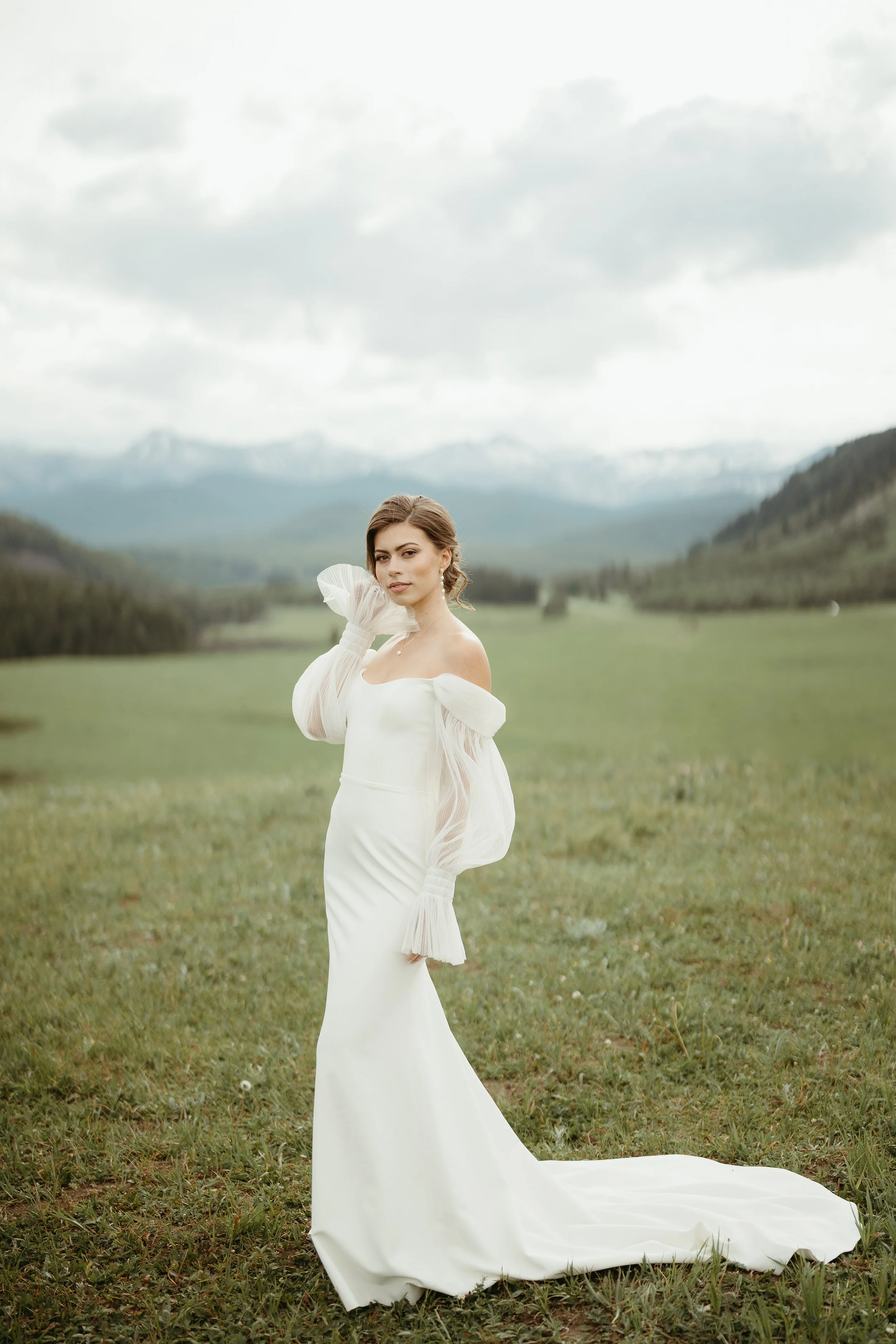 A bride stands in a field looking into the camera while being surrounded by mountains. June 2025 Kananaskis Elopement/Wedding Session with KC Photography