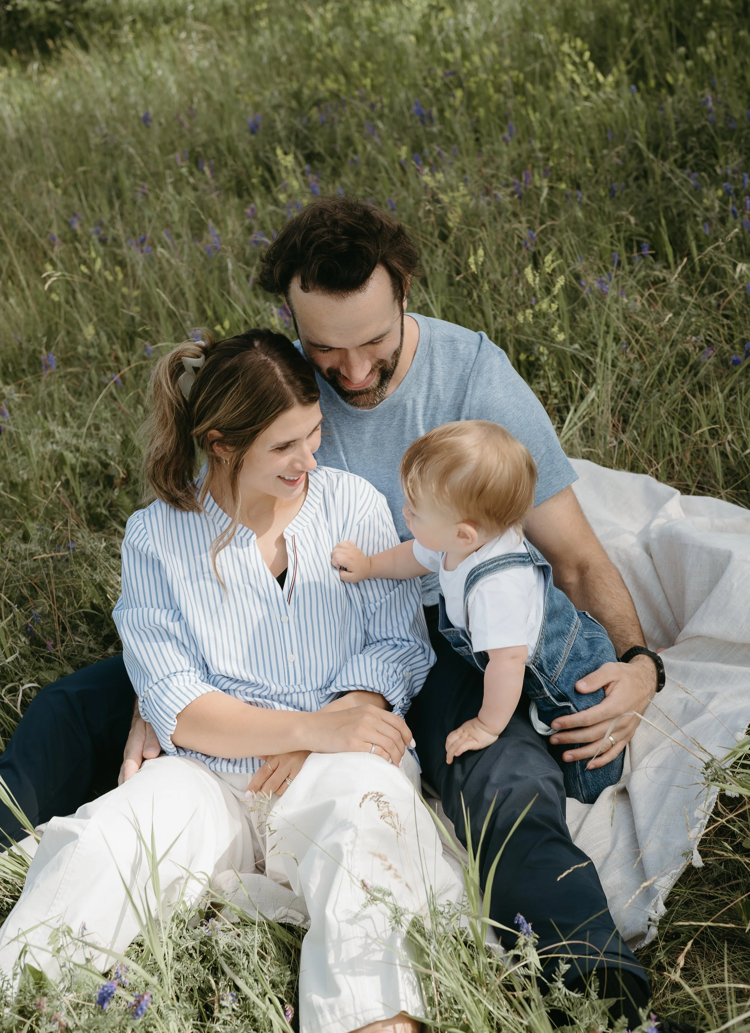 A mother sits in the lap of her husband while looking at their infant who holds onto their legs, attempting to stand in a field. July 2025 Calgary Lifestyle/Family Session with KC Photography