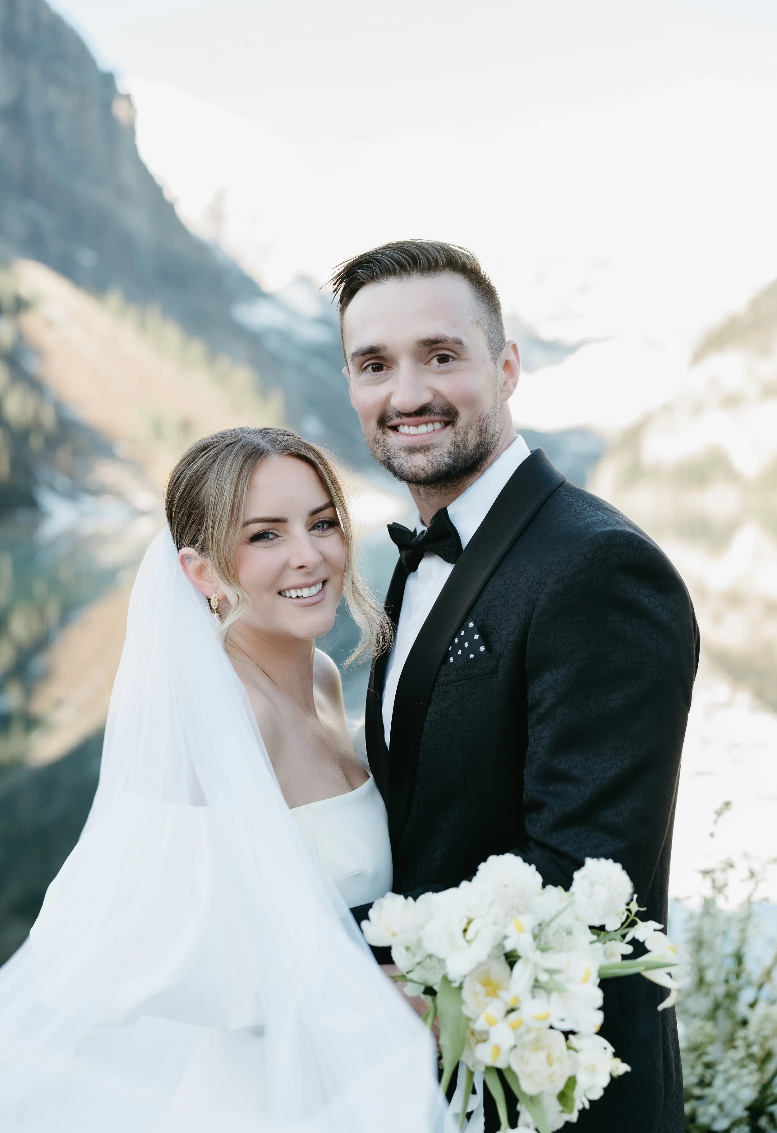 A bride and groom smile into the camera with a lake and mountains in the background. July 2025 Banff Elopement/Wedding Session with KC Photography