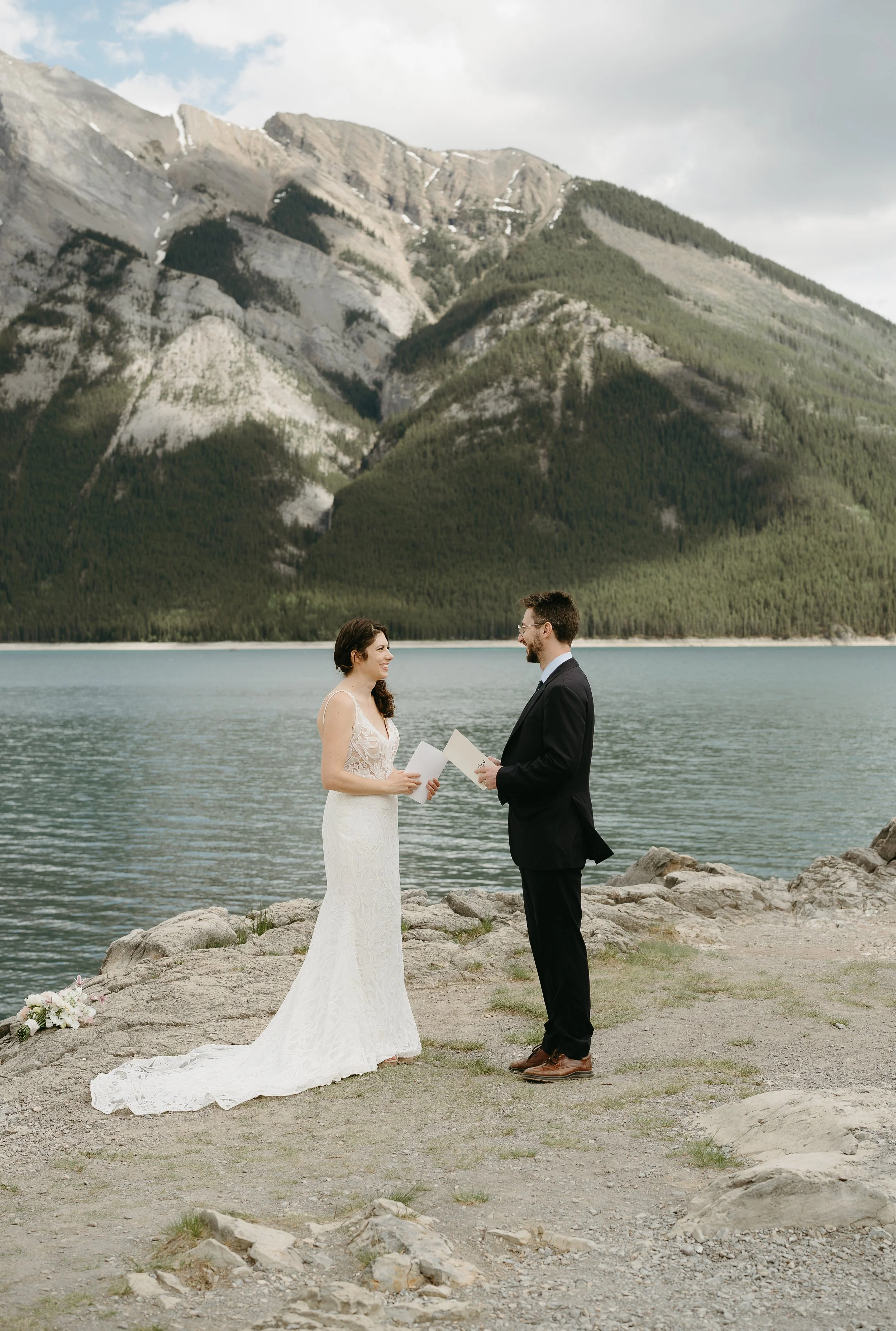 A bride and groom face one another exchanging vows on the edge of a lake surrounded by mountains. July 2025 Banff Elopement/Wedding Session with KC Photography
