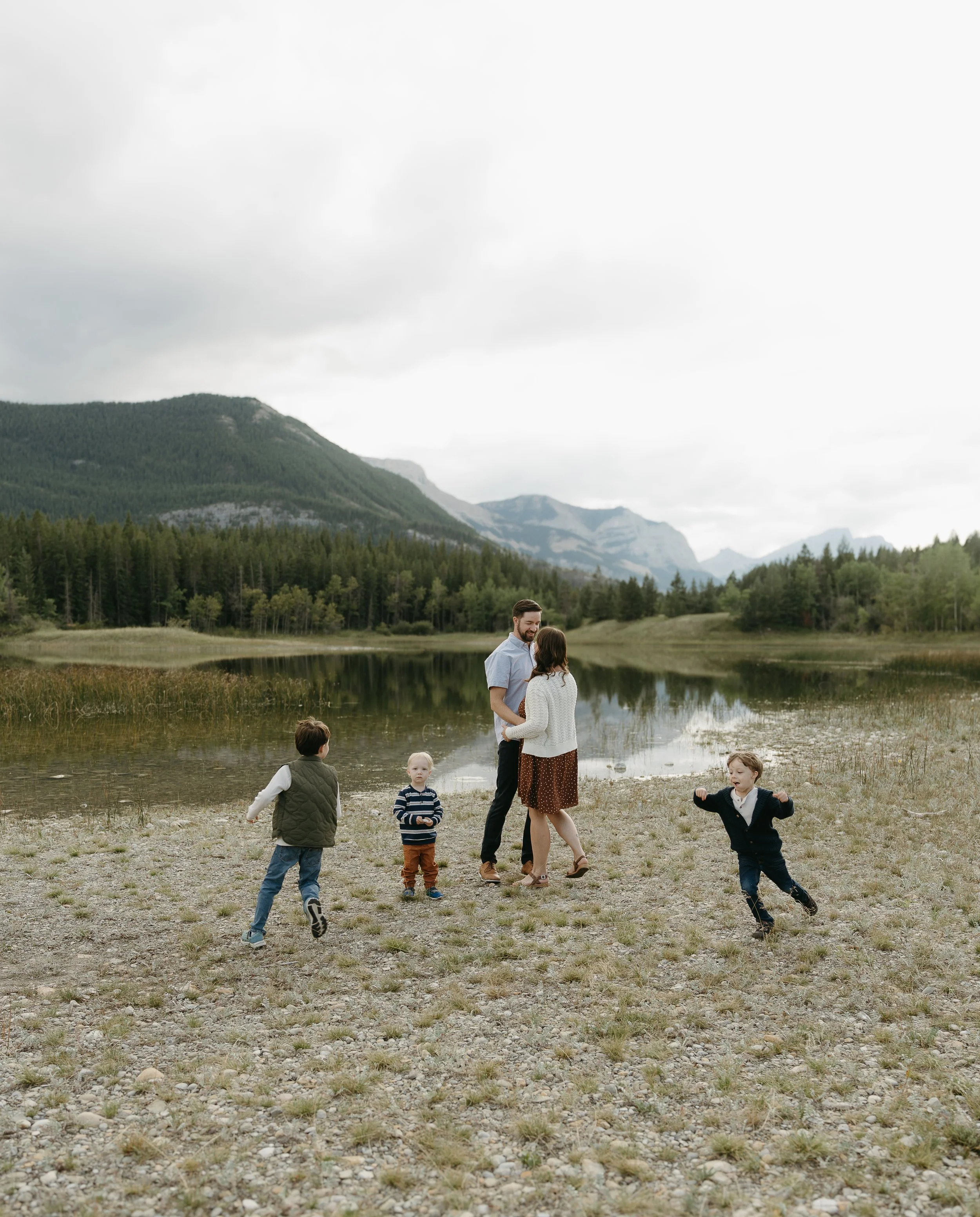 A couple holds each other in a field beside a creek while their three toddlers circle around them. Calgary Lifestyle/Family Session with KC Photography
