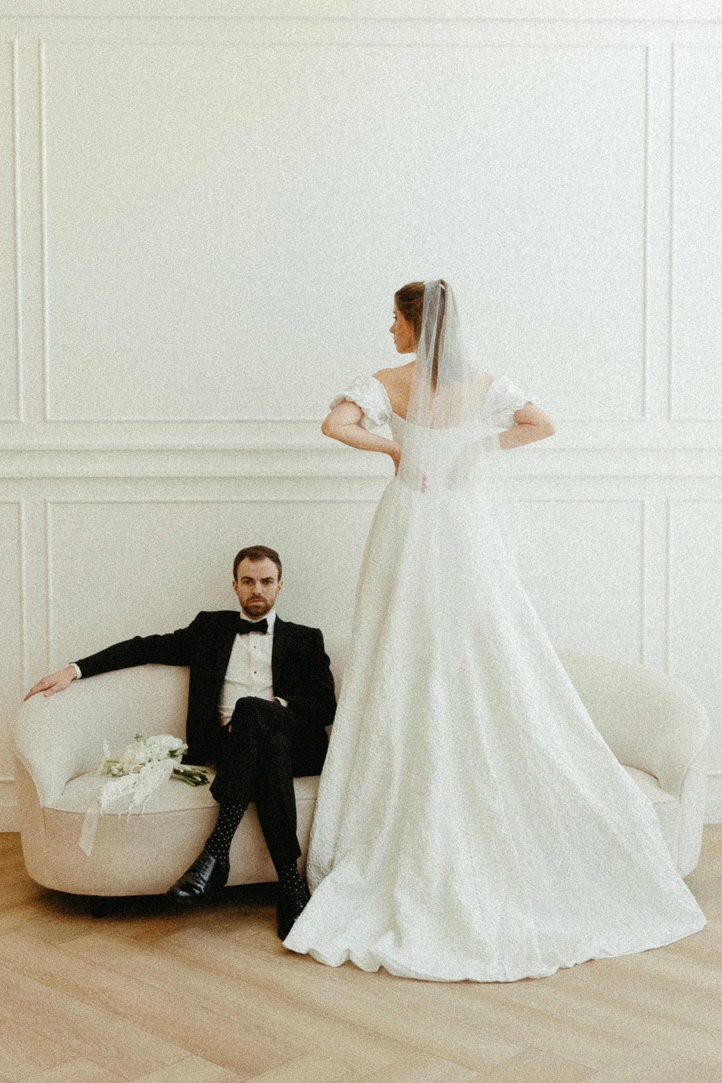 A groom sits on a couch with a wedding bouquet while his bride stands in front with her back to the camera. Calgary Elopement/Wedding Editorial Session with KC Photography