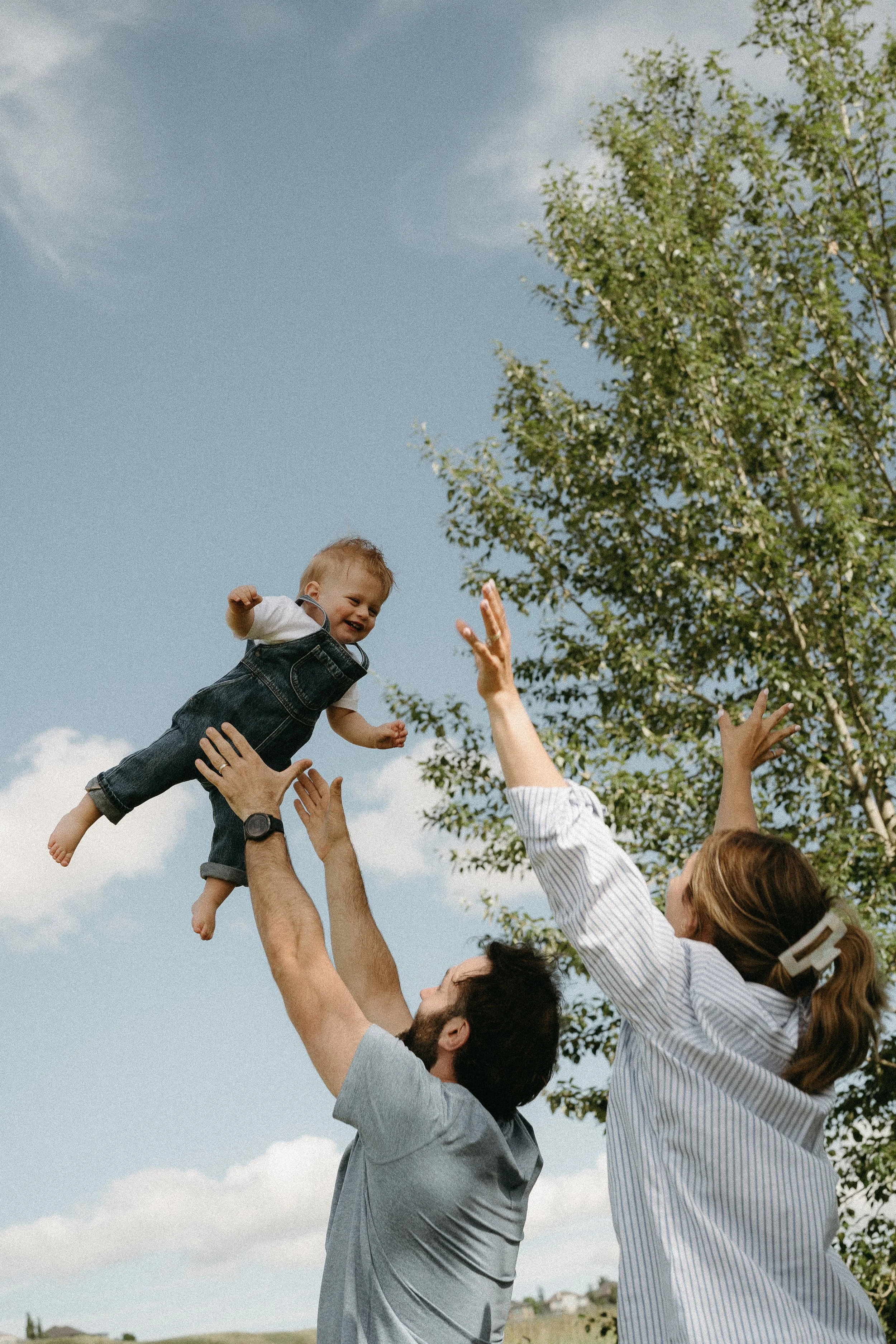 A couple playfully tosses their infant in the blue sky while the infant laughs. July 2025 Calgary Lifestyle/Family Session with KC Photography