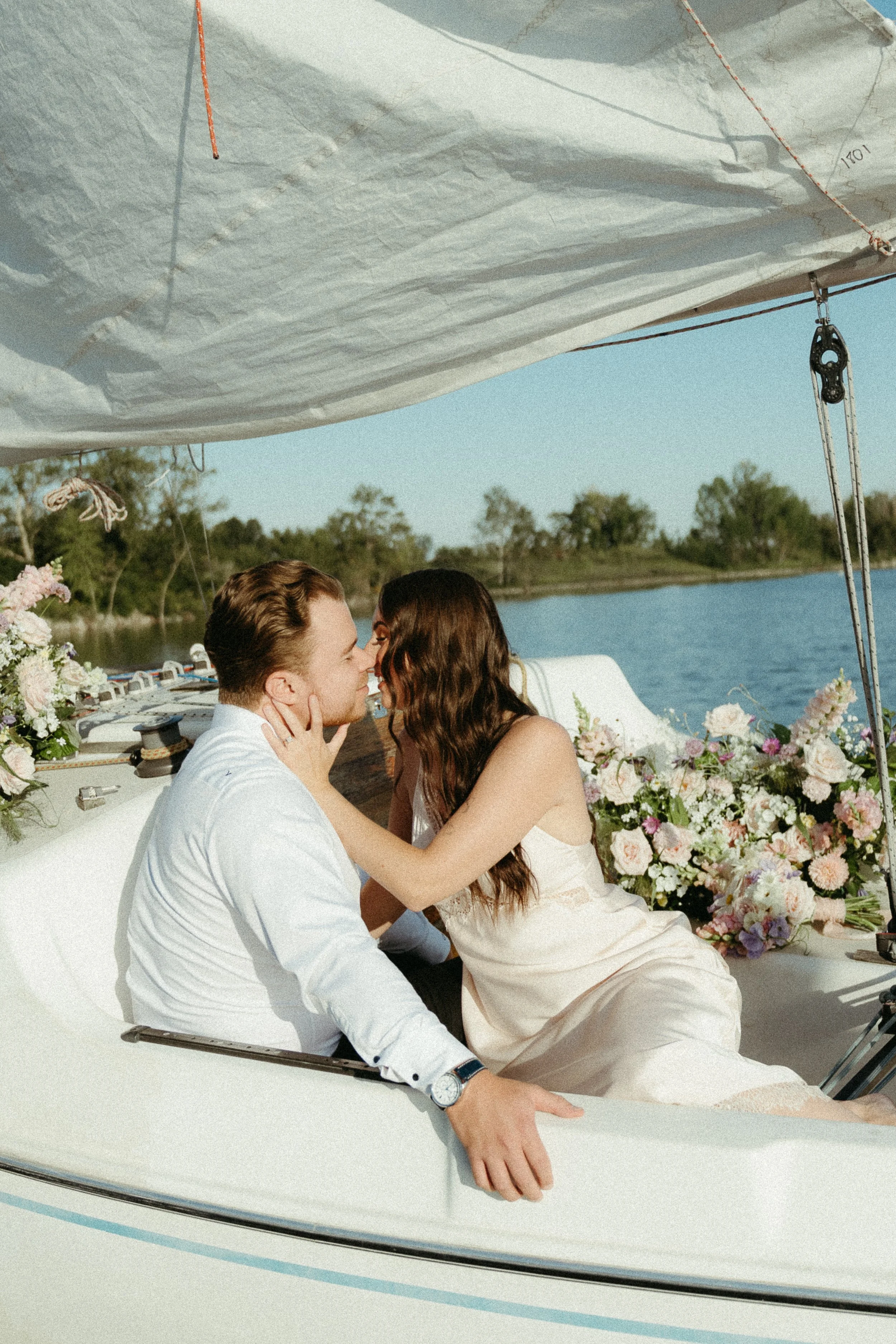 A bride leans in to kiss her groom as they sit on a sailboat covered with bouquets of flowers on the water of a lake. August 2025 Elopement/Wedding Sailboat/Sailing Session with KC Photography