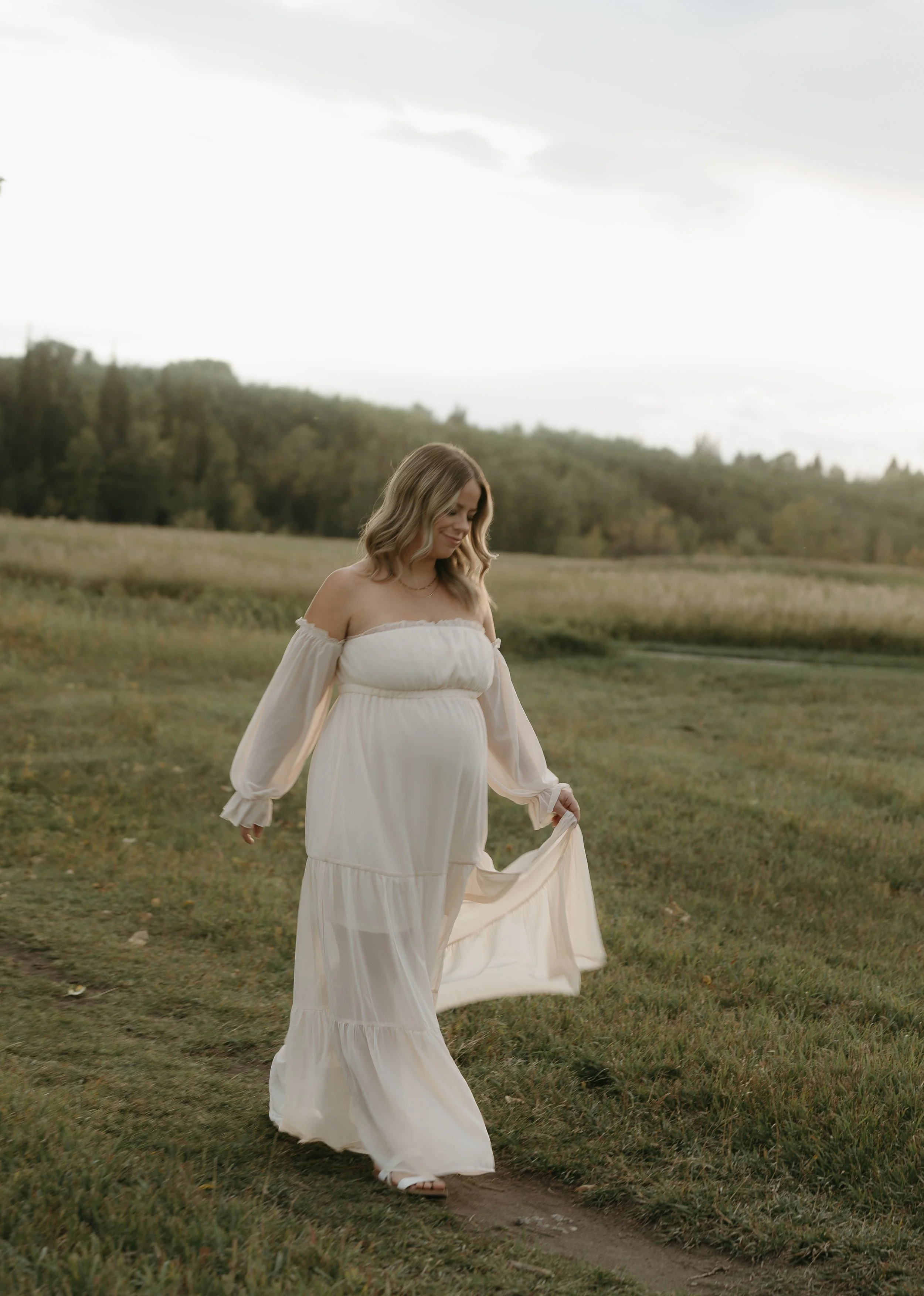 A pregnant woman walks along a dirt pathway in a field holding her dress. Kananaskis Lifestyle/Couple/Pregnancy/Maternity Session with KC Photography