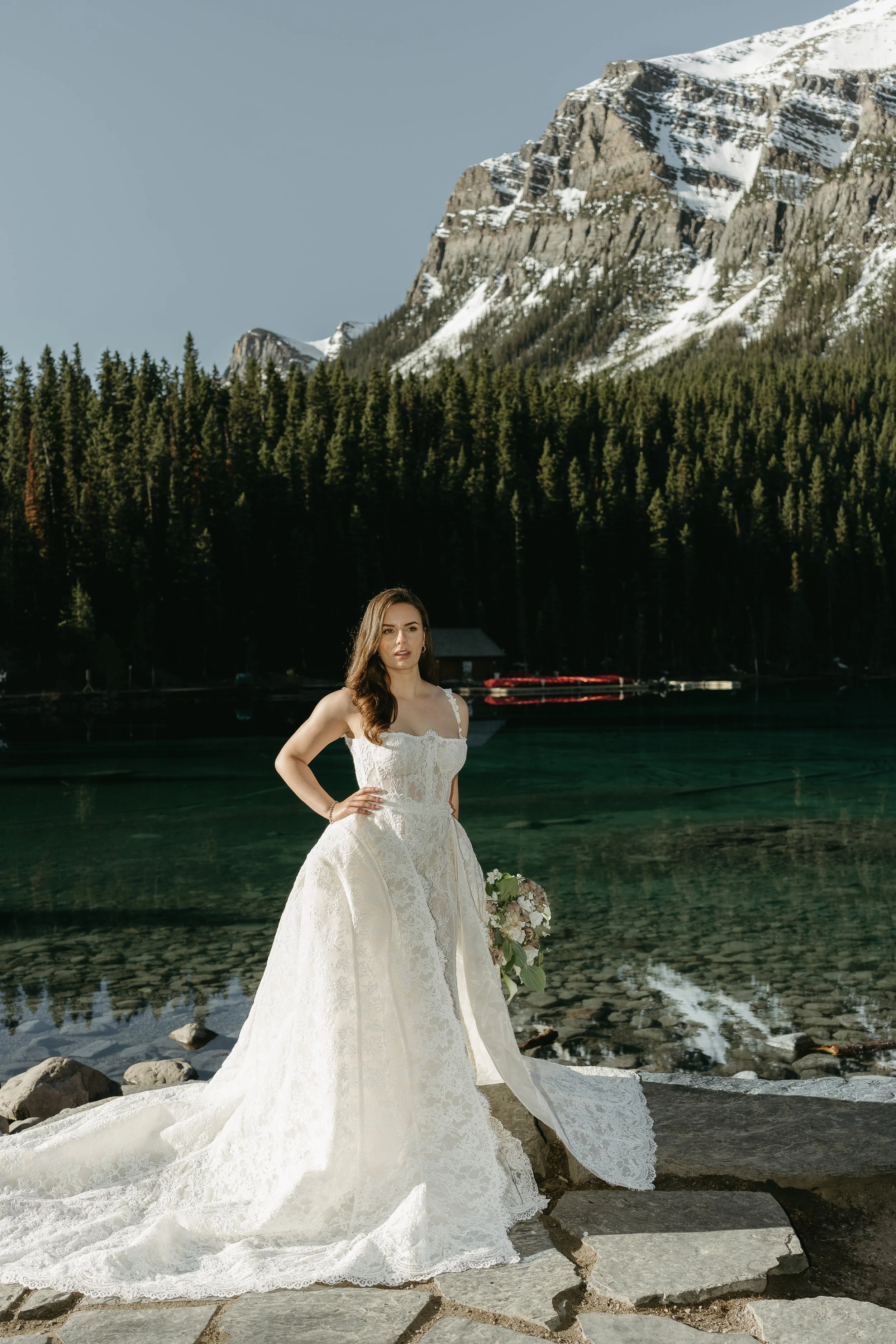 A bride stands on the edge of Lake Louise with a mountain pictured in the background. June 2025 Lake Louise Elopement/Wedding Editorial Session with KC Photography